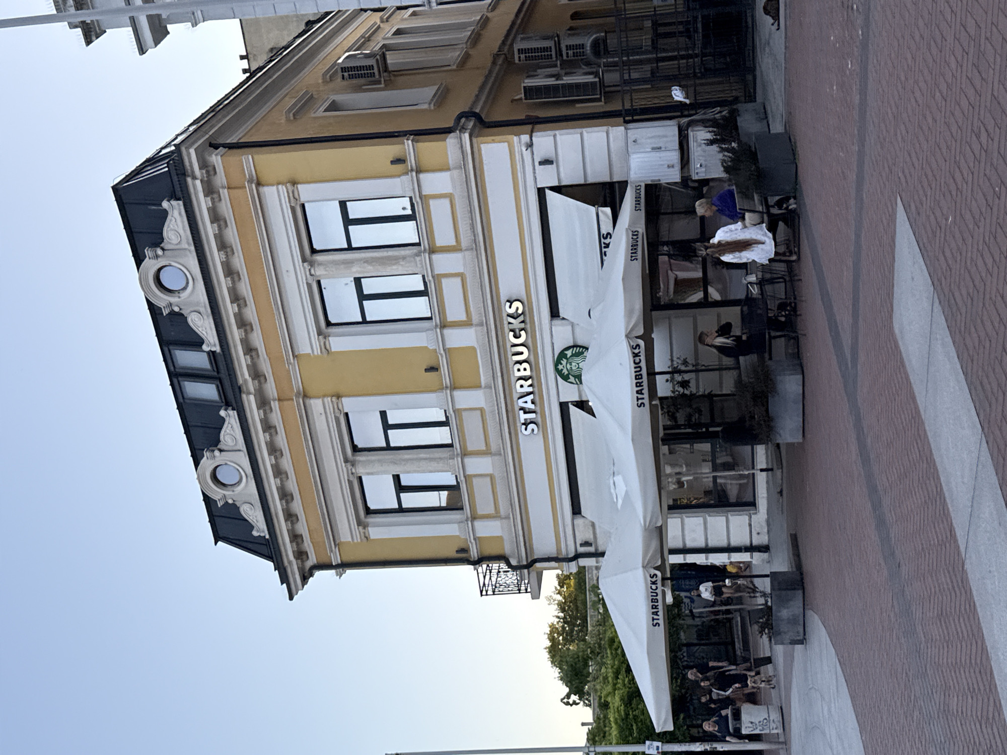 A Starbucks is visible on the ground floor of a yellow, ornate building. White umbrellas with the Starbucks logo are set up outside, providing shade over tables. Several people are seated at tables, either indoors or outdoors. The building has multiple windows and a black roof with decorative elements. The ground in front of the building is paved with red bricks. The sky is clear and blue.