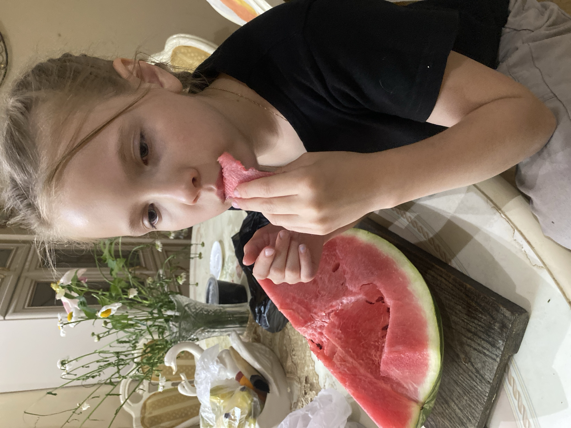 A young girl with blonde hair is eating a slice of watermelon in a room. She is wearing a black t-shirt and grey shorts. A large slice of watermelon is on a wooden board on the table in front of her. The table is covered with a patterned tablecloth. In the background, there is a vase of flowers, a white swan-shaped dish, and some chairs. The room appears to be a dining room or living area, possibly in a home in the city of Volzhsk, Russia.
