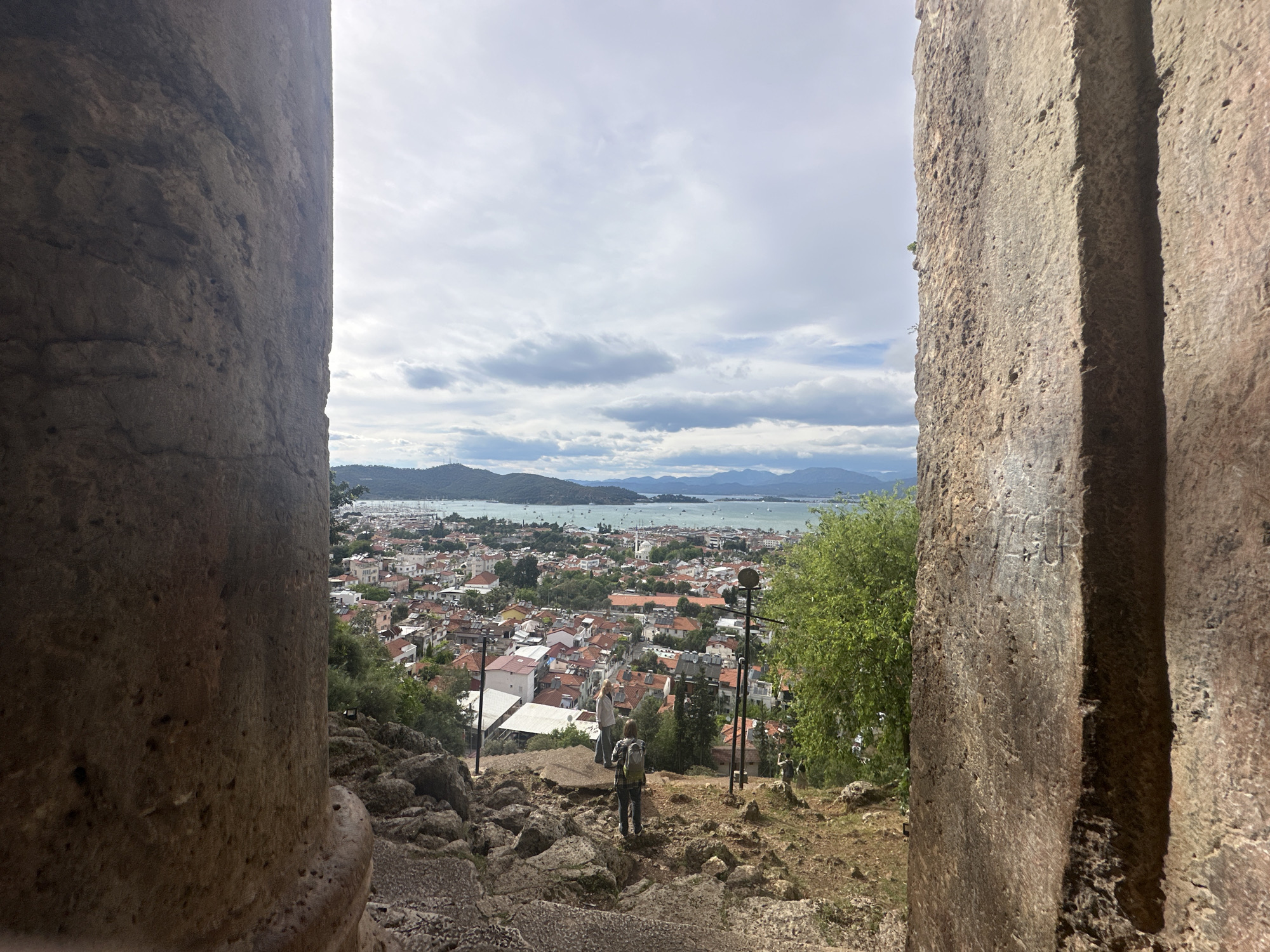 The image presents a view of Fethiye, Turkey, as seen through a stone opening, likely part of an ancient structure. The foreground is dominated by the rough, weathered stone of the opening, with one side appearing more like a rounded column and the other side featuring angular grooves. The view beyond shows a cityscape with numerous buildings topped with red roofs, interspersed with greenery. In the distance, a body of water is visible, possibly the Mediterranean Sea, with hills and mountains lining the horizon under a cloudy sky. A person wearing a backpack is walking down the steps, contributing to the sense of depth and scale within the scene. Electric poles are visible among the houses. The structure might be the Fethiye Amyntas Rock Tombs.