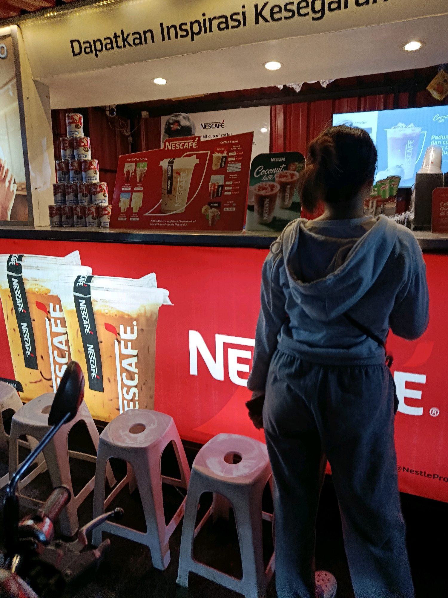 The image depicts an outdoor stall, likely a coffee kiosk, in Bekasi, Indonesia. A young woman wearing a grey hoodie and sweatpants stands with her back to the camera, facing the stall. The stall features prominent red branding with the Nescafé logo. Large posters display images of iced Nescafé coffee drinks. Canned Nescafé products are stacked in the background. Several white plastic stools are arranged in front of the stall, and a motorcycle is partially visible on the left side of the frame. The background shows a red corrugated metal wall and a screen displaying Nescafé advertisements. A sign above the stall reads "Dapatkan Inspirasi Kesegaran" which translates to "Get Fresh Inspiration." The overall scene suggests a commercial setting for a beverage vendor.