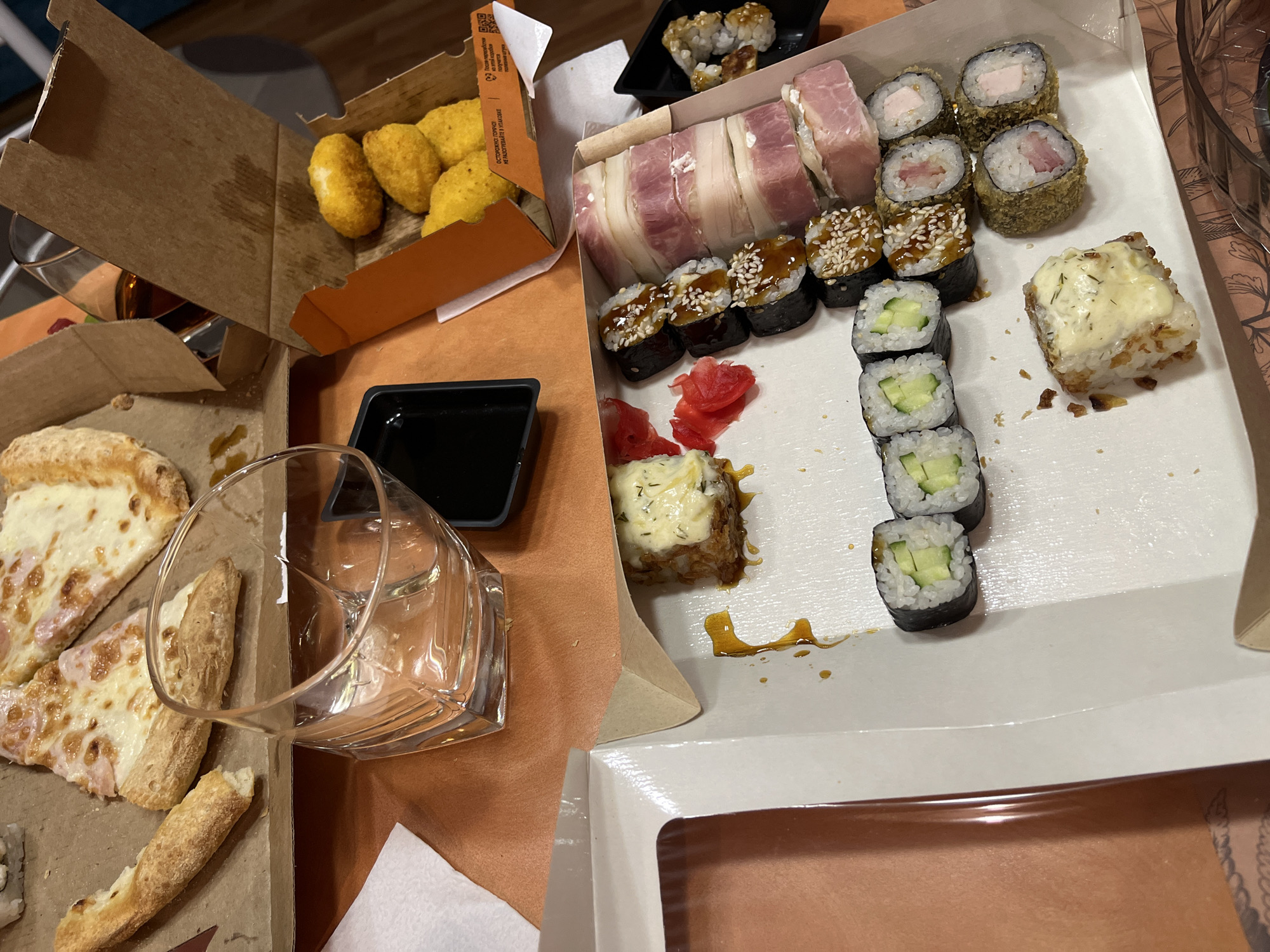 The image depicts an assortment of food in takeout containers, arranged on what appears to be a table. There's an open box of pizza slices, another with what looks like fried cheese bites, and a rectangular container holding various types of sushi rolls, including those wrapped in bacon and ones with cucumber filling. A small square container of dark sauce, likely soy sauce, is also present. A glass of water is visible near the pizza box. The photograph was taken in Chaikovsky, Russia.