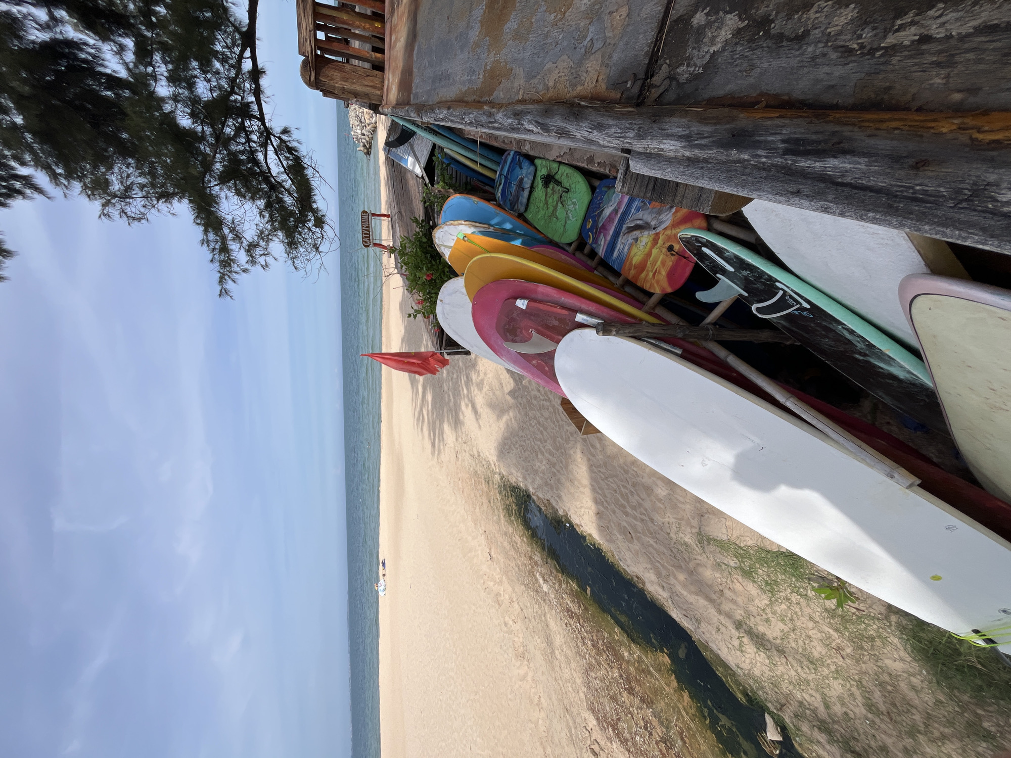 The image depicts a beach scene in Phu Thuy Ward, Vietnam. A row of surfboards of various colors and designs are neatly stacked on the sand, leaning against a wooden structure. In the background, the ocean stretches to the horizon under a clear blue sky with scattered clouds. The beach itself is sandy, with a small channel of dark water running through it, possibly a stream or a drainage ditch. A red flag is planted on the sand near the water's edge. The overall impression is one of a seaside recreational area, likely a surf rental shop or a resort.