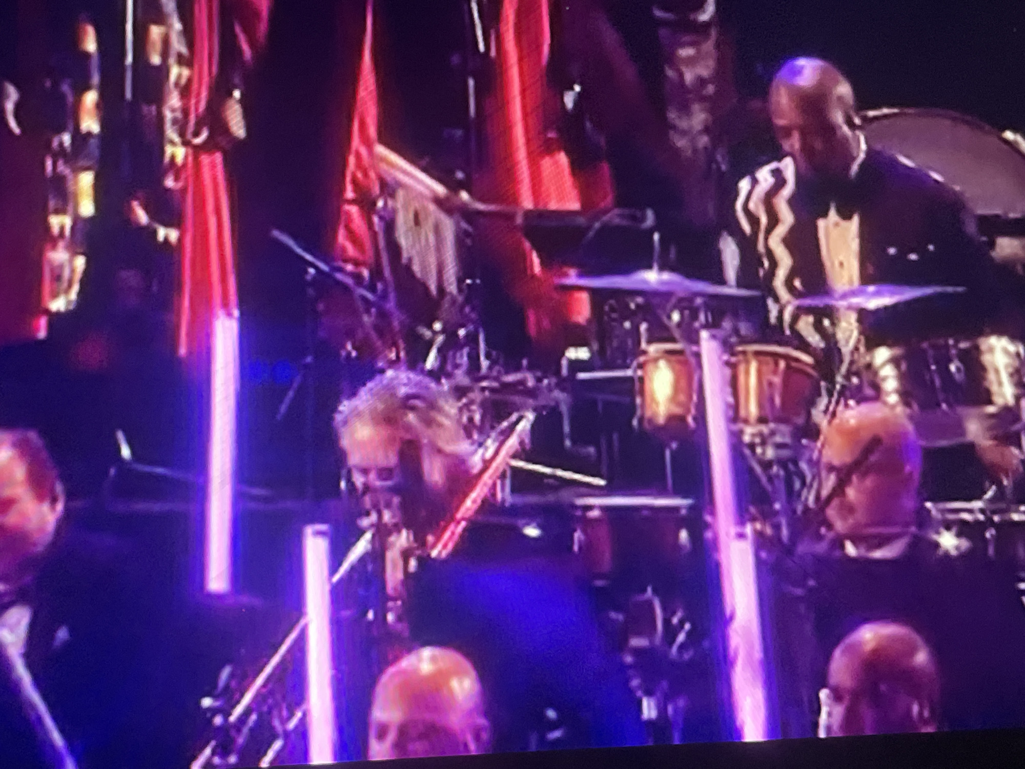 A group of musicians are performing on a stage. In the foreground, a drummer with blond hair is playing drums and cymbals. He is wearing a dark blue suit. To his right, another drummer with a shaved head and glasses is playing drums. He is wearing a black suit. In the background, a bald man in a black tuxedo is playing drums. He is wearing a black and white striped shirt. The stage is decorated with red fabric and illuminated by purple lights. There are several drum sets and other musical instruments on the stage. The audience is not visible.