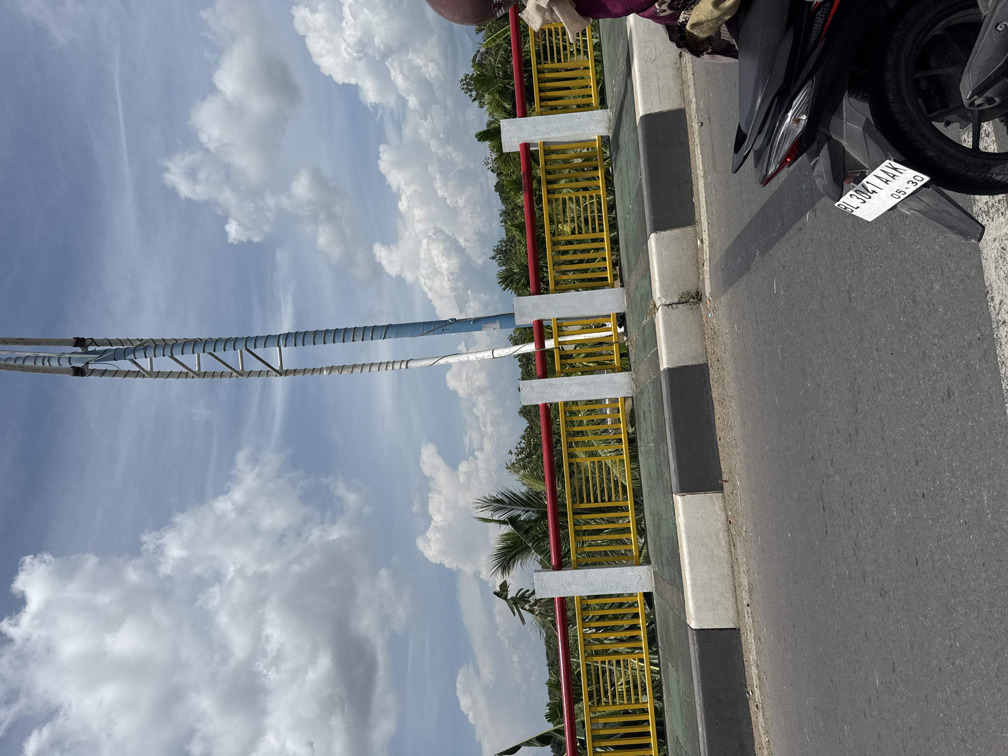 In Lambaro, Indonesia, a street view captures a bright, overcast day. The foreground features the back of a black motorcycle with license plate BL 3041 AAK, suggesting it is parked or stopped near the edge of the road. The motorcycle is partially obscured by the camera's perspective, but its taillight and rear wheel are visible. Beyond the motorcycle, a road surface is evident, marked by a concrete curb with black and white segments.
The middle ground is dominated by a bridge or overpass with a bright yellow metal railing, topped by a red bar. White pillars support the railing at intervals. Behind the railing, lush green foliage, including palm trees, creates a natural backdrop. A tall, thin structure, possibly a telecommunications tower or utility pole, with a blue and white segmented shaft, stands prominently against the sky.
The sky is filled with large, fluffy white clouds against a light blue canvas, indicating a dynamic weather pattern. The overall scene depicts a typical day in an Indonesian town, with infrastructure and natural elements coexisting.