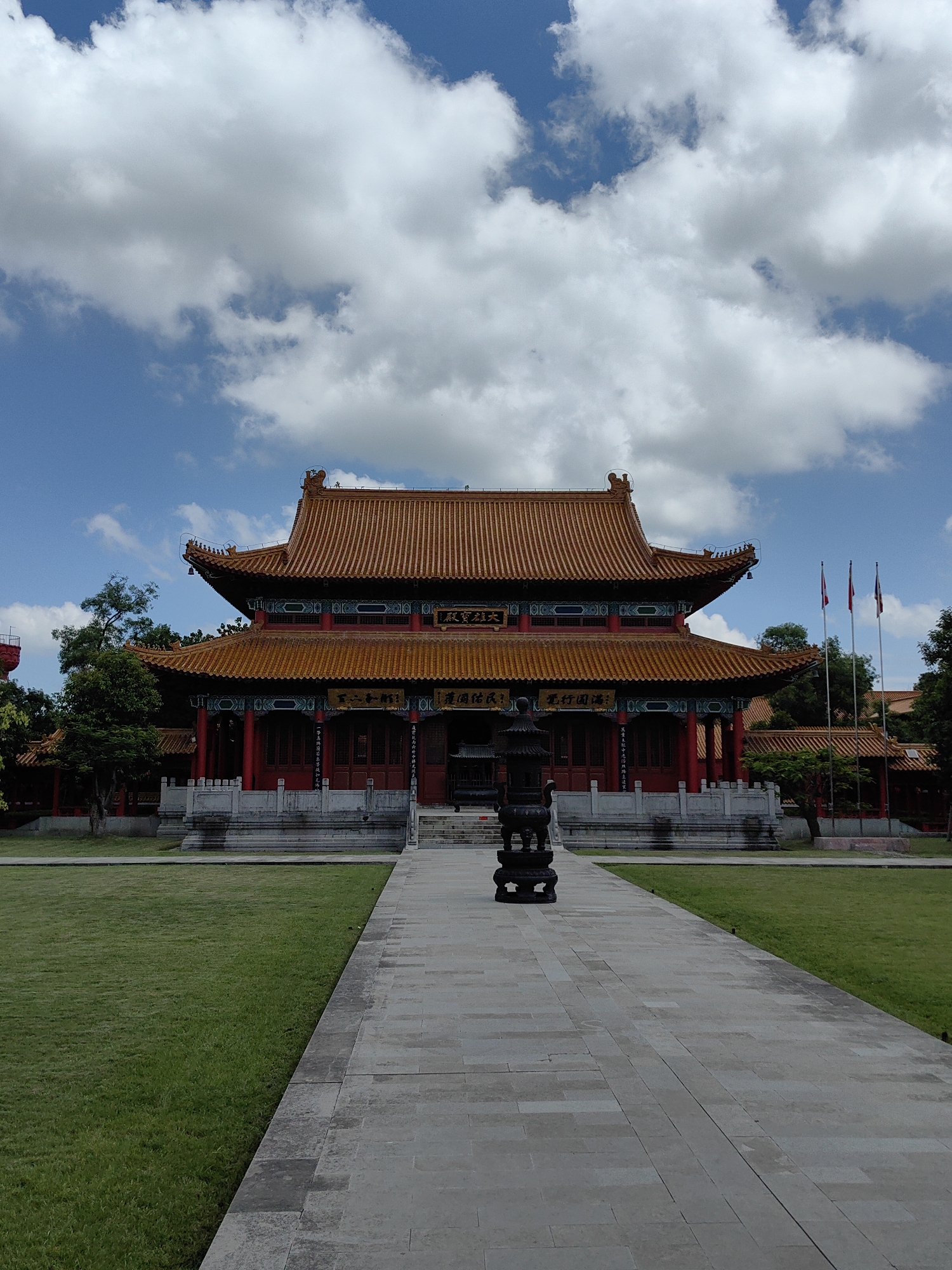 A medium shot captures the front facade of a large, traditional East Asian temple, likely Buddhist, under a partly cloudy sky. The temple features a prominent, orange-tiled roof with intricate upturned eaves and decorative elements. The building's lower sections are painted red with ornate architectural details. A wide, paved walkway leads towards the temple entrance, flanked by a neatly manicured green lawn on the left. In the foreground, on the walkway, stands a dark, ornate incense burner. Several flags on poles are visible to the right of the temple. The overall scene is serene and well-maintained, suggesting a place of worship or cultural significance. The location is identified as Lumbini Bazaar, Nepal.