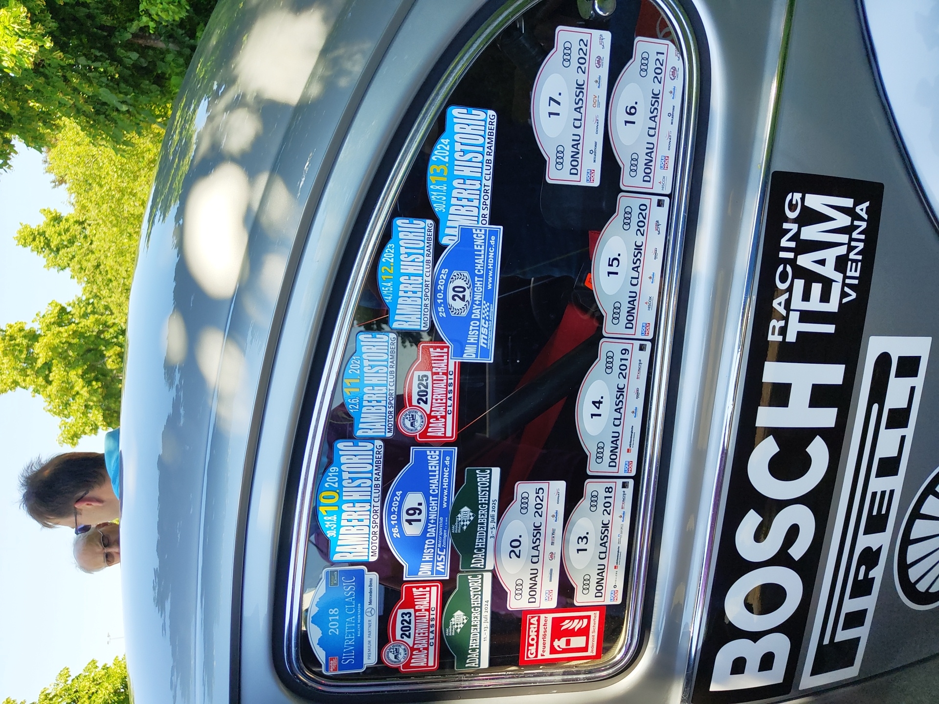 The image shows the rear passenger side window of a silver car, adorned with numerous stickers for various car rallies and events. The stickers include "Silvretta Classic 2018," "Ramberg Historic" (multiple years), "ADAC-Bayerwald-Rallye" (multiple years), "DMI Histo Day+Night Challenge," and "Donau Classic" (multiple years). A "Gloria Feuerlöscher" (fire extinguisher) sticker is also visible. The car is parked outdoors, with bright sunlight casting reflections on its surface. Green trees and a bright blue sky are visible in the background. Two men are partially visible through the window, looking into the car. One man has gray hair and is wearing glasses, while the other has dark hair and is wearing sunglasses. Below the window, a large "BOSCH" sticker and a "PIRELLI" sticker are prominently displayed on the car's bodywork, along with a "RACING TEAM VIENNA" sticker. The image appears to have been taken in Pirmasens, Germany, as indicated by a sticker reading "Pirmasens" on one of the rally decals.