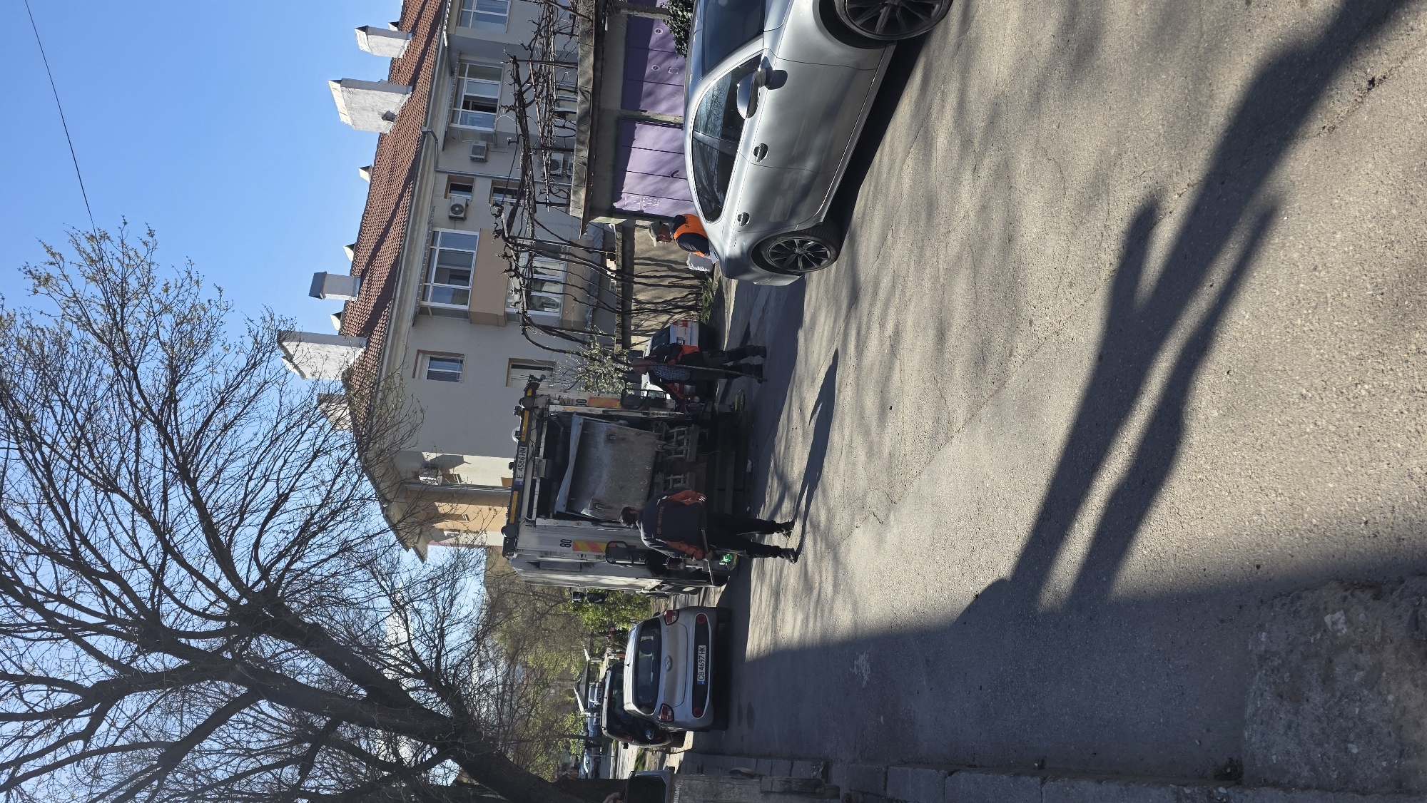 The image shows a street in Blagoevgrad, Bulgaria. A garbage truck is parked on the street, with several sanitation workers nearby. A silver car is parked in front of the truck and another silver car is parked behind the truck. A multi-story building is on the left side of the frame. There is a bare tree in the lower left corner of the frame. The sky is blue and clear. The sun casts shadows on the street. The license plate of one of the cars is visible.