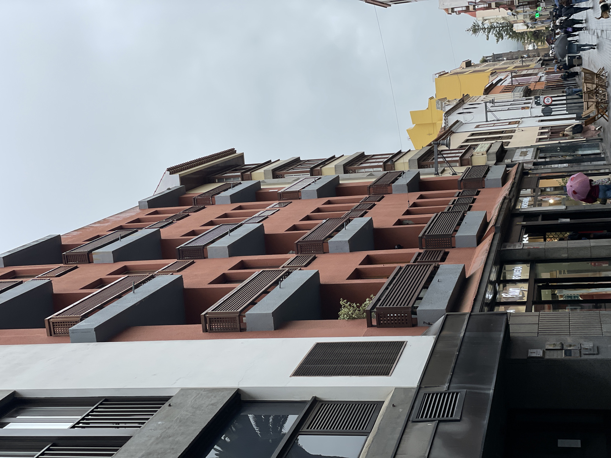 The image shows a street view in San Cristóbal De La Laguna, Spain, focusing on a modern building facade. The building is characterized by a reddish-brown exterior with projecting, angular gray balconies and wooden lattice screens. The lower portion of the building is white with rectangular ventilation grates. The building rises up and along the right edge a street is seen with people walking under umbrellas. Various shops with large glass windows are visible, and the facades of other buildings line the street. The sky is overcast.