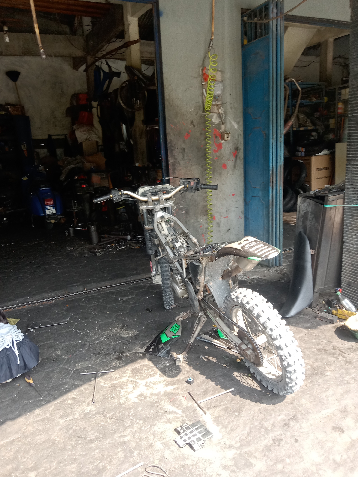 The image depicts the interior of a motorcycle repair shop in Siwalan, Indonesia, bathed in sunlight. In the foreground, a disassembled dirt bike is the primary subject, with its frame, wheels, and various parts scattered around. Tools, including a T-handle socket wrench and a screwdriver, are visible on the concrete floor, indicating ongoing repair work. A blue scooter is parked in the background, to the left of the frame. The shop environment appears cluttered with motorcycle parts, shelves stocked with items, and a large blue folding door, partially open, revealing more of the shop's interior. The walls of the shop are concrete, and overhead, wooden beams support the roof structure. The overall scene suggests an active but somewhat disorganized workspace typical of a small automotive repair business. There are no people visible in the image.
