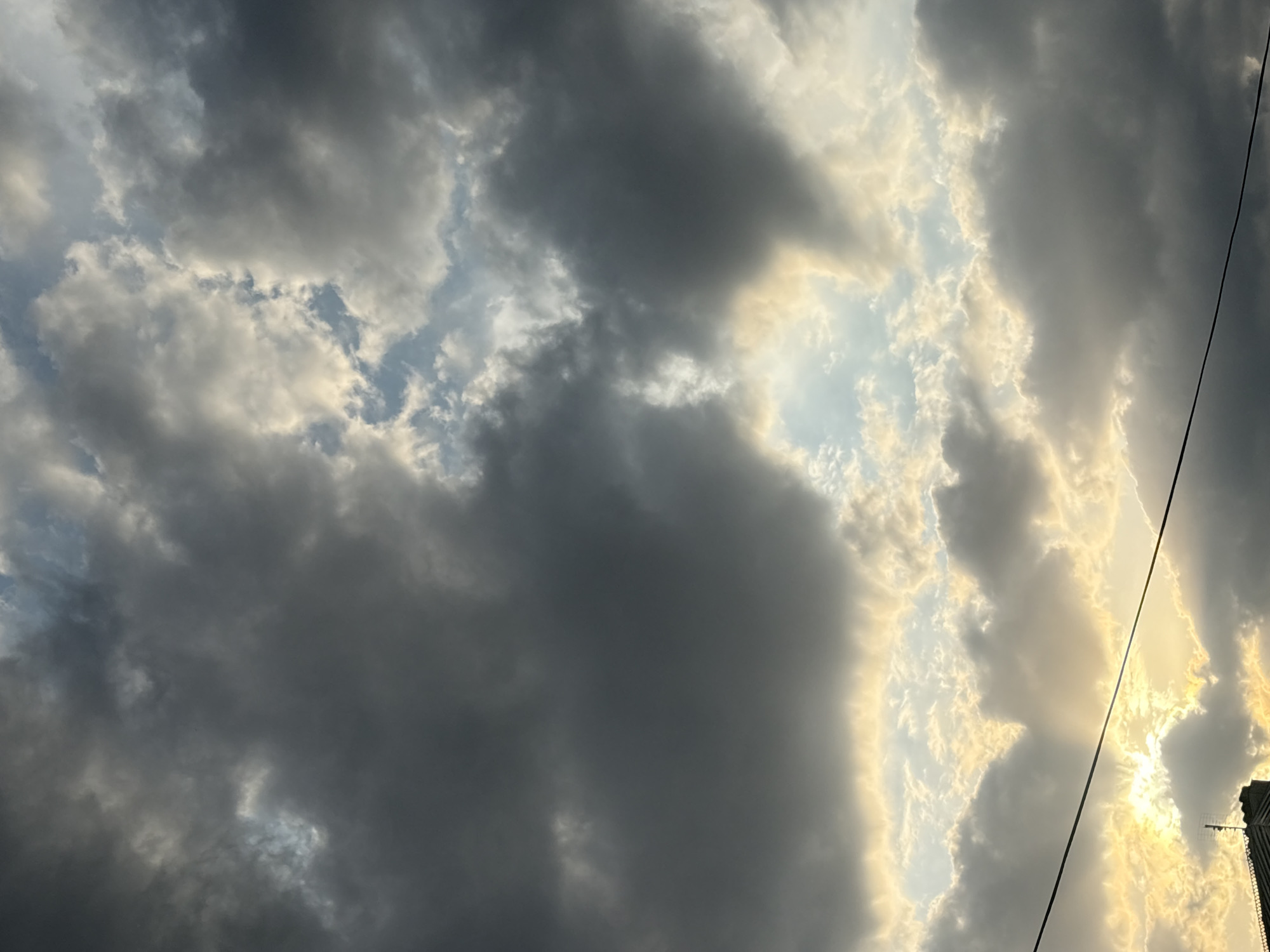 The image shows a dramatic sky filled with dark, heavy clouds illuminated by the setting sun. Rays of light break through gaps in the clouds, creating a strong contrast between light and shadow. In the lower left corner, the silhouette of a building with a spire and a visible power line extending across the frame indicate an urban setting. The location is specified as Georgiyevsk, Russia. There are no people or discernible actions in the image; it is a still scene focused on the atmospheric conditions.