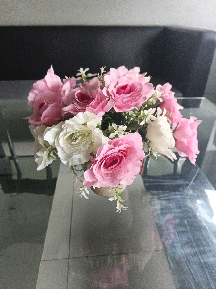 The image displays a close-up, eye-level view of an artificial flower arrangement situated on a reflective glass-top table within an indoor setting. The bouquet primarily consists of light pink and white faux roses, interspersed with small white filler flowers and green artificial foliage. The flowers are arranged in a cylindrical pot, which appears to be covered with a textured, light brown material, possibly burlap or a similar fabric.
The glass surface of the table reflects the base of the pot, the lower parts of the flowers, and parts of the room's environment, including the tiled floor below the table. The flooring visible beneath the table features light-colored, rectangular tiles. In the blurred background, a dark, indistinct object, likely a piece of furniture such as a sofa or cabinet, occupies the upper portion of the frame. The lighting suggests an interior space. No individuals, actions, or interactions are depicted. While the prompt specifies the location as the Special Capital Region of Jakarta, Indonesia, no discernible geographical markers within the image itself confirm this specific location.