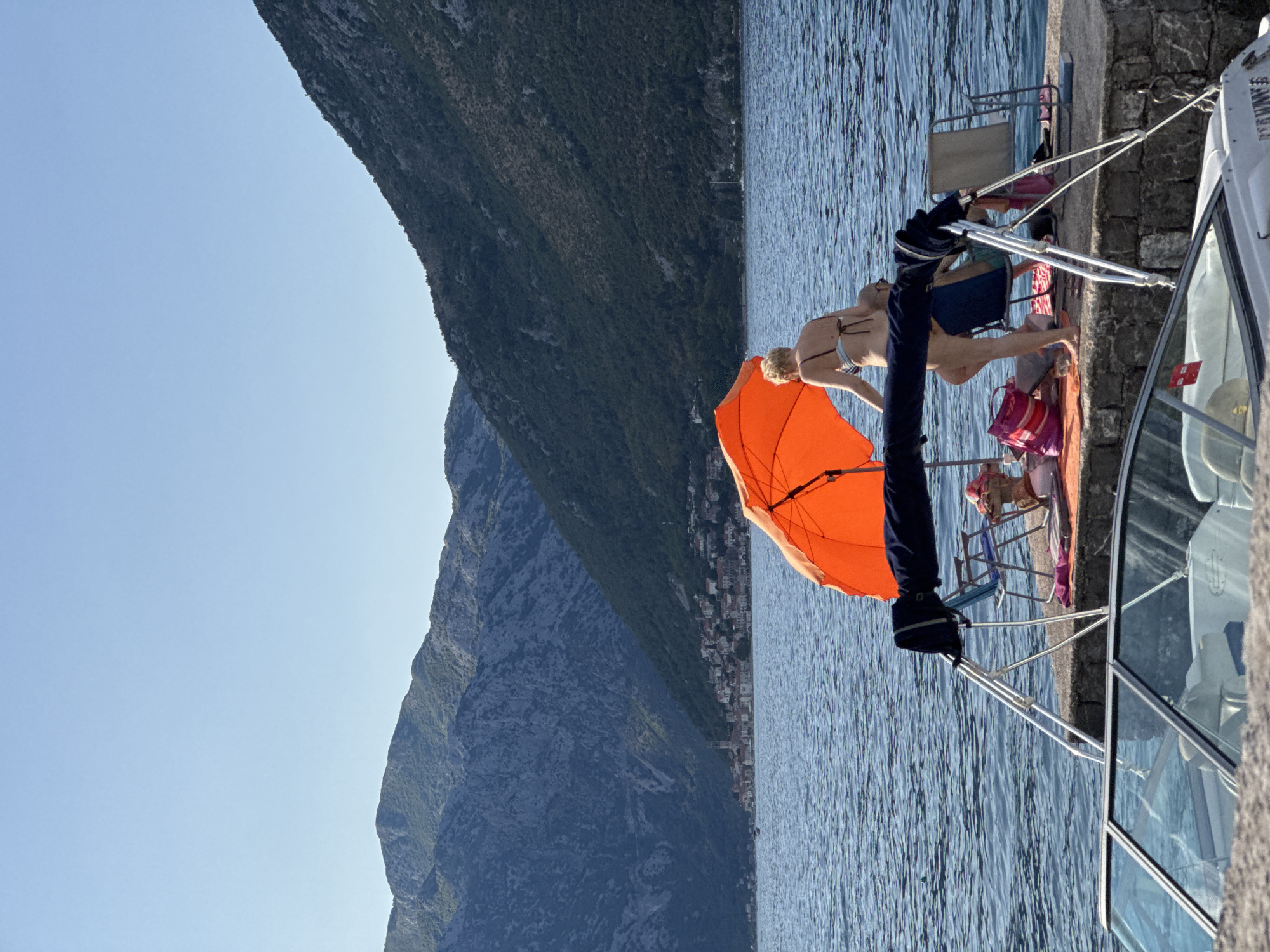 In the city of Perast, Montenegro, a woman in a bikini stands near an open orange umbrella on a paved waterfront. Behind her, a man sits in a blue chair. Other beach items, including bags and towels, are scattered on the ground. The water is calm and blue, with a boat docked in the foreground. The background features lush green mountains and a town with a bell tower.