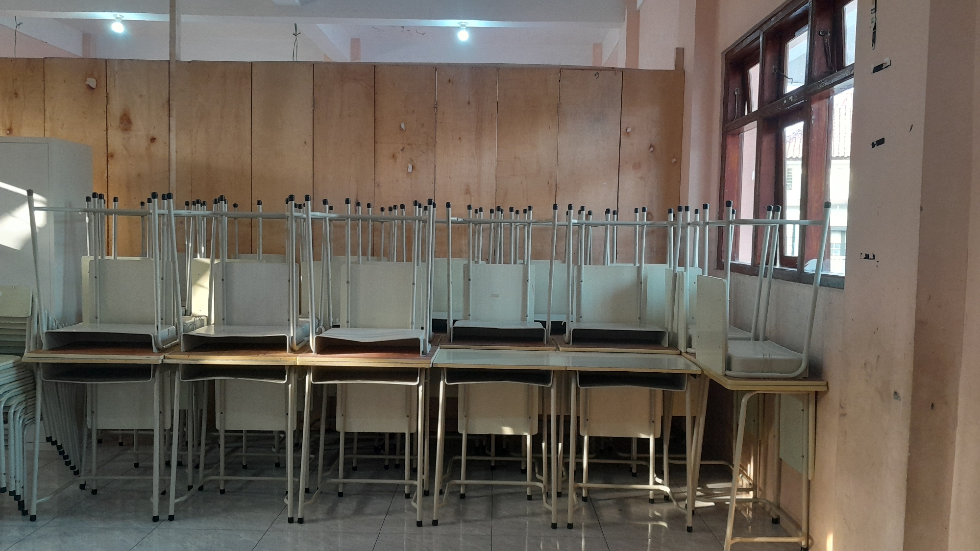 The image depicts an empty classroom in Tasikmalaya, Indonesia. The room is sparsely furnished with a series of identical desks, each with a chair stacked on top. The desks are arranged in rows, facing a wall covered in light-colored wood paneling. Sunlight streams in from a window on the right side of the room, casting long shadows across the floor and desks. The overall impression is one of stillness and abandonment, suggesting the room is not in use. The scene is forensic in its detail, capturing the precise arrangement of the furniture and the play of light and shadow without interpretation.