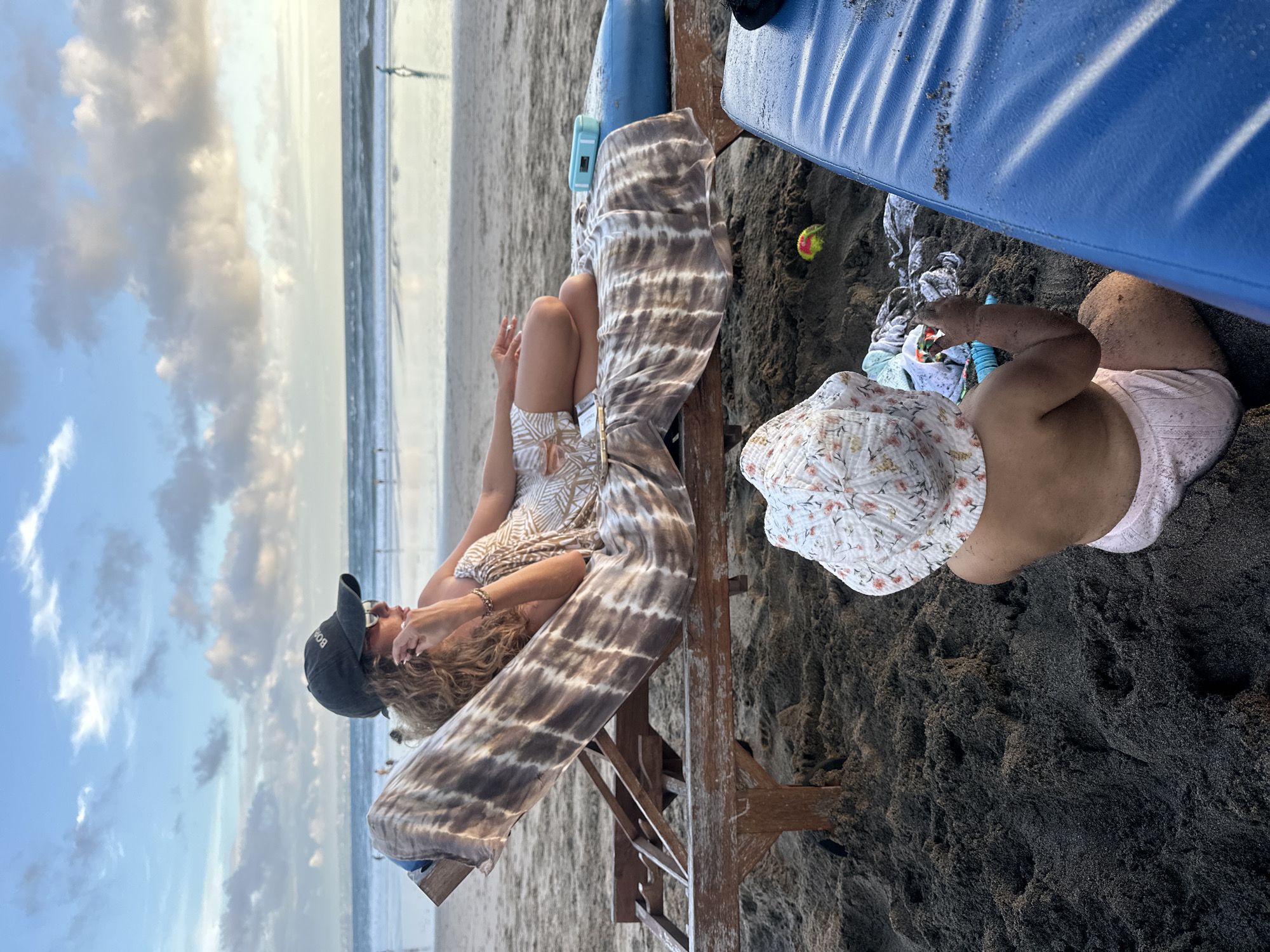 The scene depicts a woman and a child on a beach in North Kuta, Indonesia. The woman is wearing a patterned romper, a black baseball cap, and sunglasses. She is sitting on a wooden structure, with a tie-dye patterned garment draped over it. The child, wearing a floral sunhat and a light-colored outfit, is playing in the black sand. There are blue objects (possibly surfboards) nearby, along with a small, multicolored ball. In the background, the sky shows a mix of blue and cloudy patches, and the ocean extends to the horizon. Other people are visible as distant figures along the shoreline.