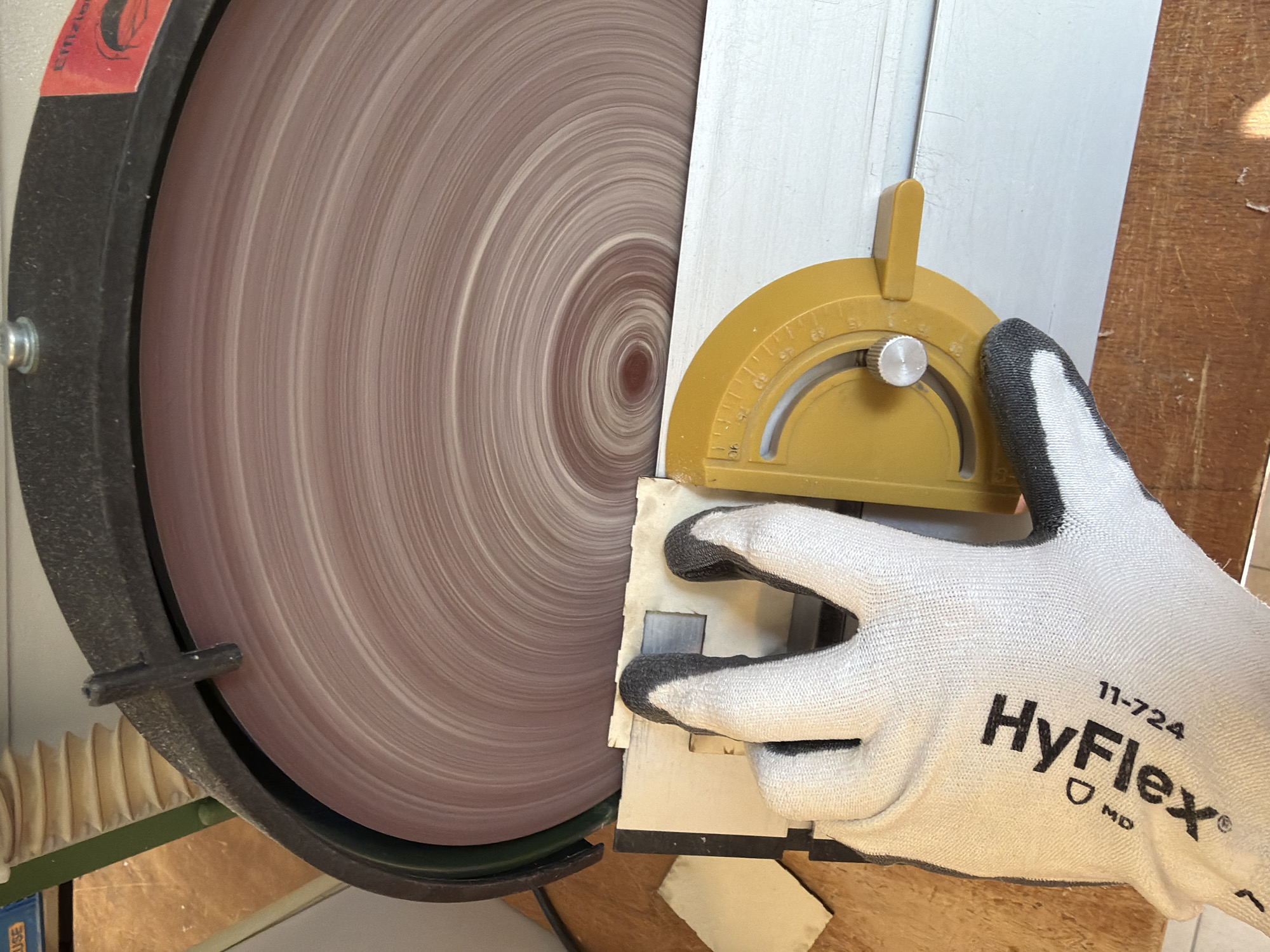 A gloved hand wearing a "HyFlex" work glove holds a piece of white material against a disc sander with a yellow protractor to measure the angle. The scene is indoors. There is no information to conclude the location is Milan, Italy.