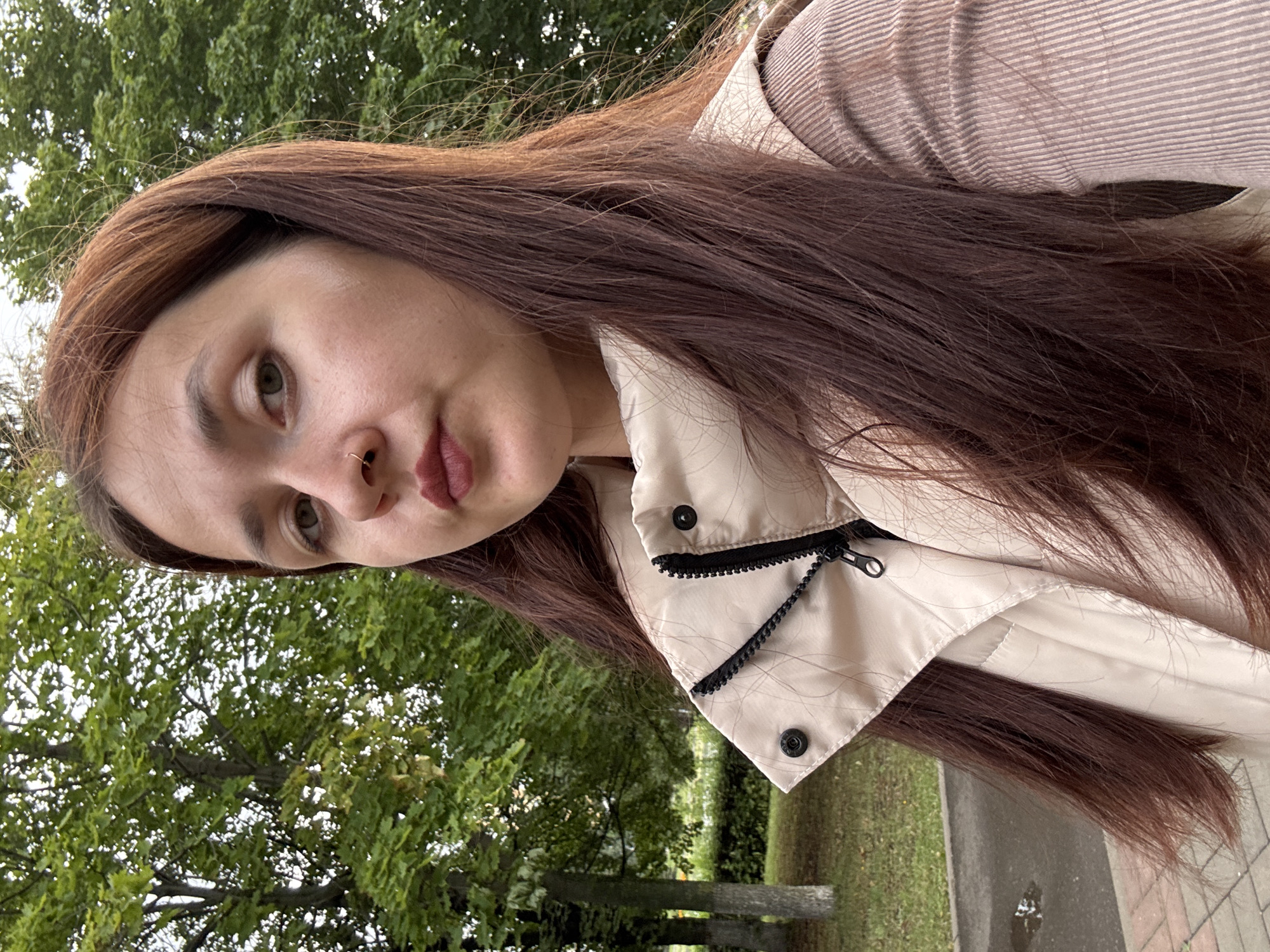 A young woman with long, brown hair and a nose ring is taking a selfie outdoors. She is wearing a beige puffer vest over a brown ribbed shirt. In the background, there are trees and a paved path. The location is identified as Naberezhnye Chelny, Russia. The lighting suggests it is daytime, possibly overcast.