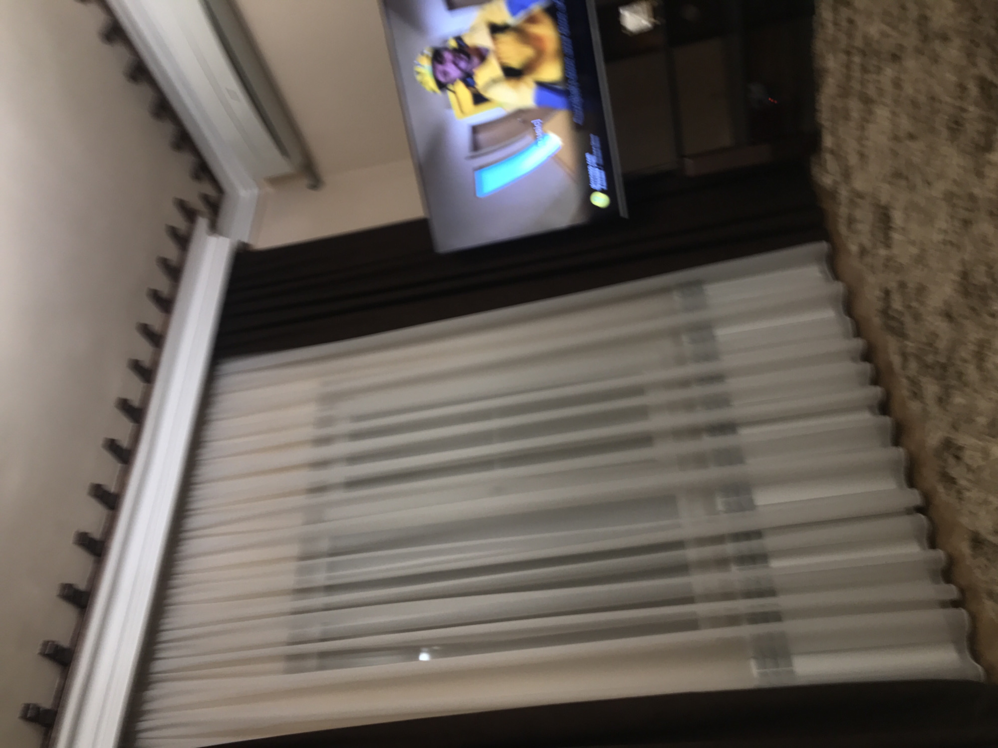 A blurred, tilted shot captures a room interior. A television displaying a person in a yellow outfit is mounted on a dark-colored wall above a window. The window is covered with white horizontal blinds. To the left of the window is a wall with a white curtain rail. To the right of the window is a textured wall. The location cannot be determined to be Yerevan, Armenia from the image.