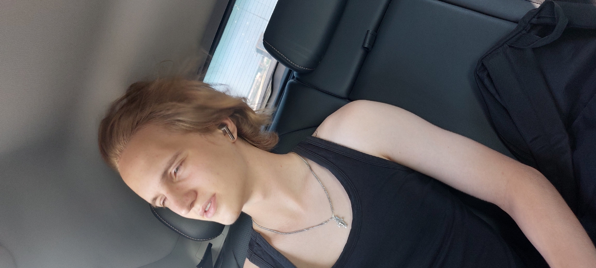 A young Caucasian male with blond hair and fair skin is seated in the back of a car, looking up and to his left with his eyes partially closed. He is wearing a black tank top and a silver chain with a cross pendant. He has a silver wireless earbud in his right ear. The interior of the car appears to be black leather. Outside the car window, a blurred cityscape is visible. The image is taken from a high angle. The location is identified as Chaikovsky, Russia.