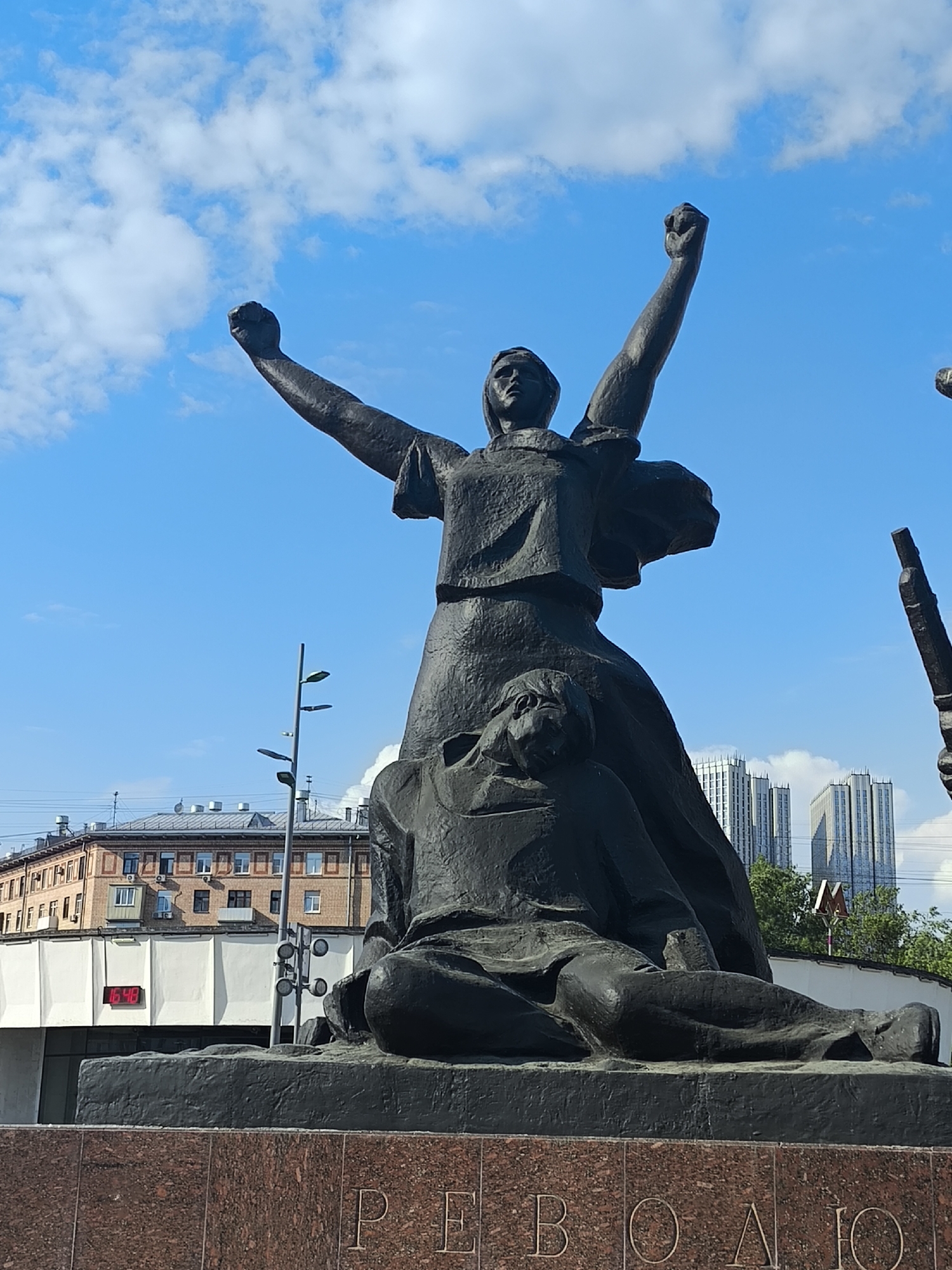 The image shows the Revolution of 1905 monument in Moscow, Russia, in front of a bright blue sky with some clouds. The bronze monument depicts a woman with both arms raised in a fist, standing over a fallen man. Behind the monument, there is a building with a red digital clock displaying "16:48". Further back, there is a street lamp, another building, and a tall modern building. A sign with an "M" logo can be seen in the background. The base of the statue has the word "РЕВОЛЮЦИЯ" (Revolution) inscribed on it.