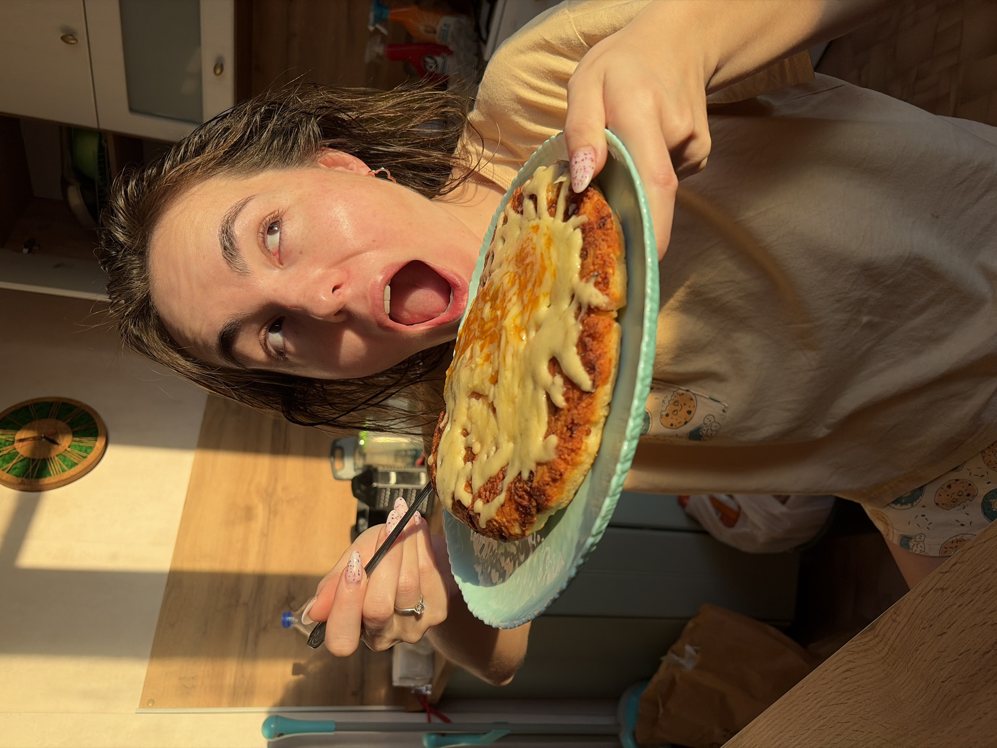 The image depicts a female individual in a sunlit domestic kitchen or dining area in Izhevsk, Russia. The subject, with dark brown, damp-looking hair, faces the camera with her mouth wide open, eyes directed upwards and to the right. She wears a light beige t-shirt and patterned shorts with cookie and donut motifs. Her left hand, adorned with a ring on the fourth finger and pink speckled nail polish, holds a dark-colored fork. Her right hand supports a light blue-green plate holding a round, baked food item covered in melted cheese and a reddish sauce.
Strong, warm-toned sunlight casts sharp shadows across the light-colored wall behind her. A round wooden wall clock with a green face and Roman numerals is mounted on this wall. To the right, white kitchen cabinets with silver handles are visible; one upper cabinet door is slightly ajar, revealing a green bowl inside. Various items are on counters or shelves in the background, including a blue-capped water bottle and two brightly colored cleaning product bottles (red and orange). A turquoise-handled cleaning tool is partially visible on the left. The lower left foreground shows a section of a light wooden table or counter, and two bags, one brown paper and one white plastic, are visible on the floor or a lower surface.