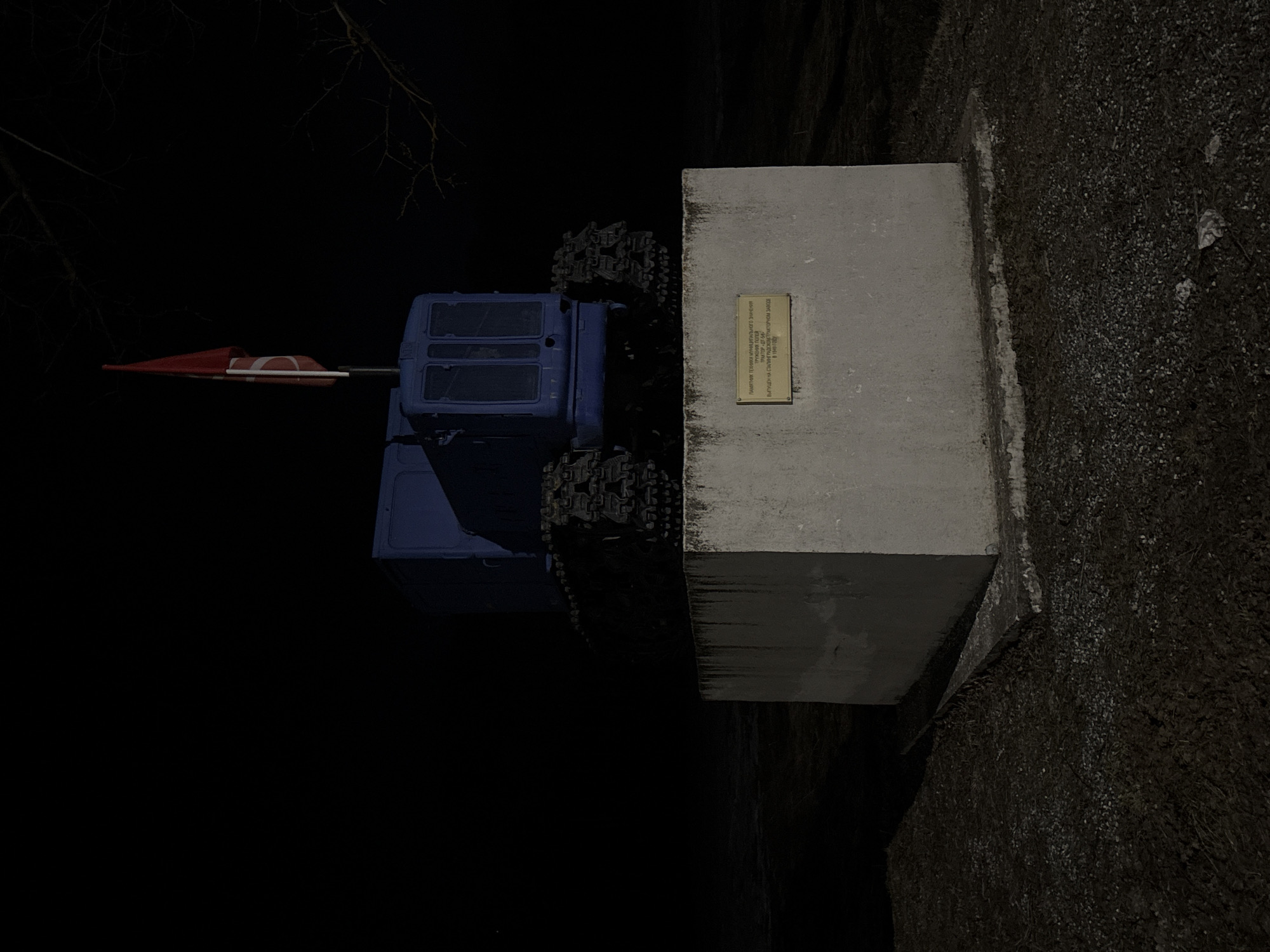 The image depicts a monument to a "Worker of the Fields" tractor, produced at the Stalingrad Tractor Plant in 1940. The tractor, painted blue, is positioned next to a concrete pedestal with a plaque. The scene is dark, possibly at night, with sparse vegetation and what appears to be a gravelly ground covering. Bare branches are visible in the upper left of the frame. According to the plaque, it is a monument of municipal significance. The location is Rusalevka, Russia.
