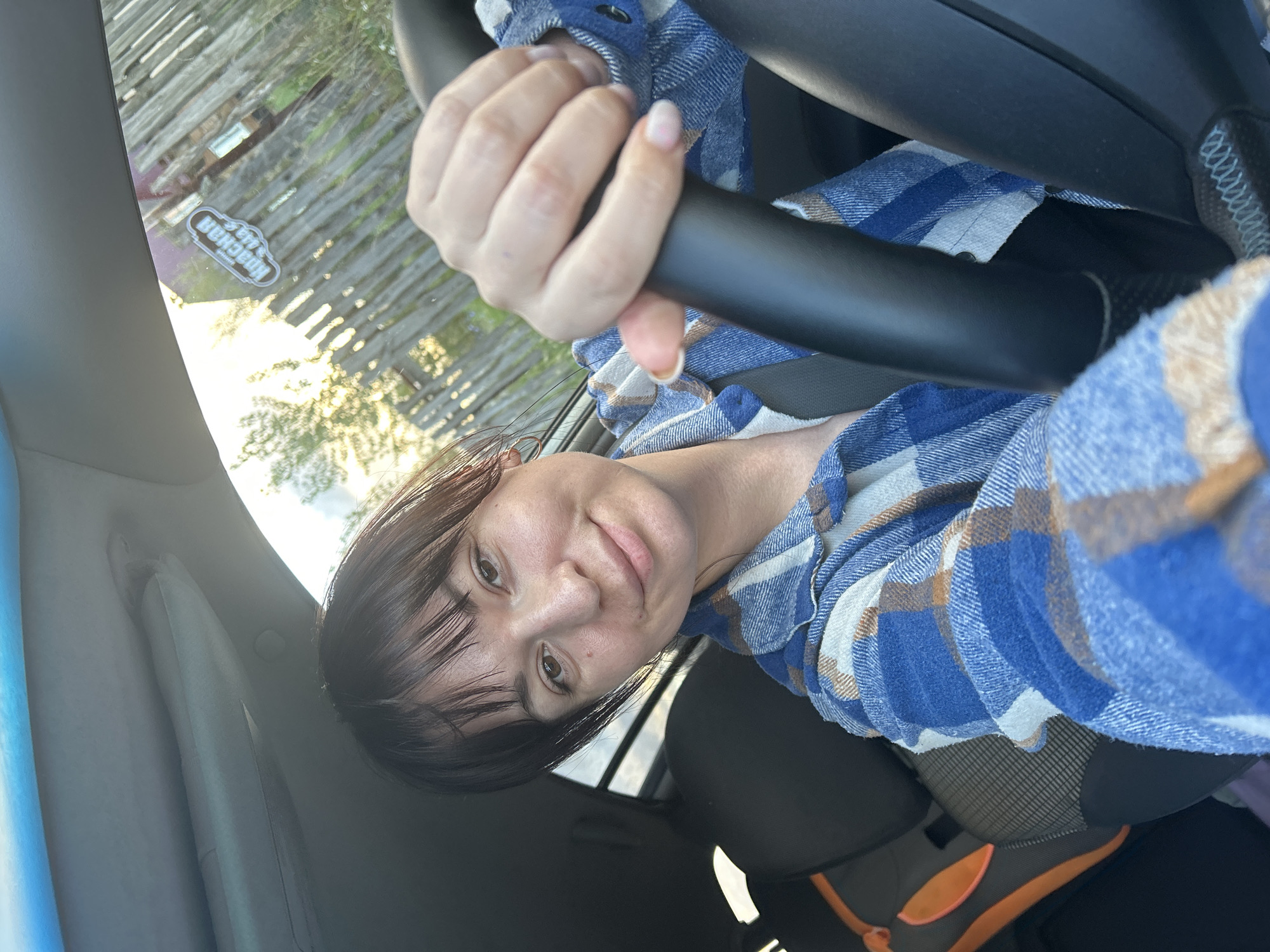 A young woman with dark hair and bangs is sitting in the driver's seat of a car, looking at the camera and smiling. She is wearing a blue and white plaid shirt. Her hands are on the steering wheel. The sun is setting, casting a warm glow through the windshield, which has a sticker on it with Russian text. Outside the car, a wooden fence is visible. The car's interior includes a car seat in the background.