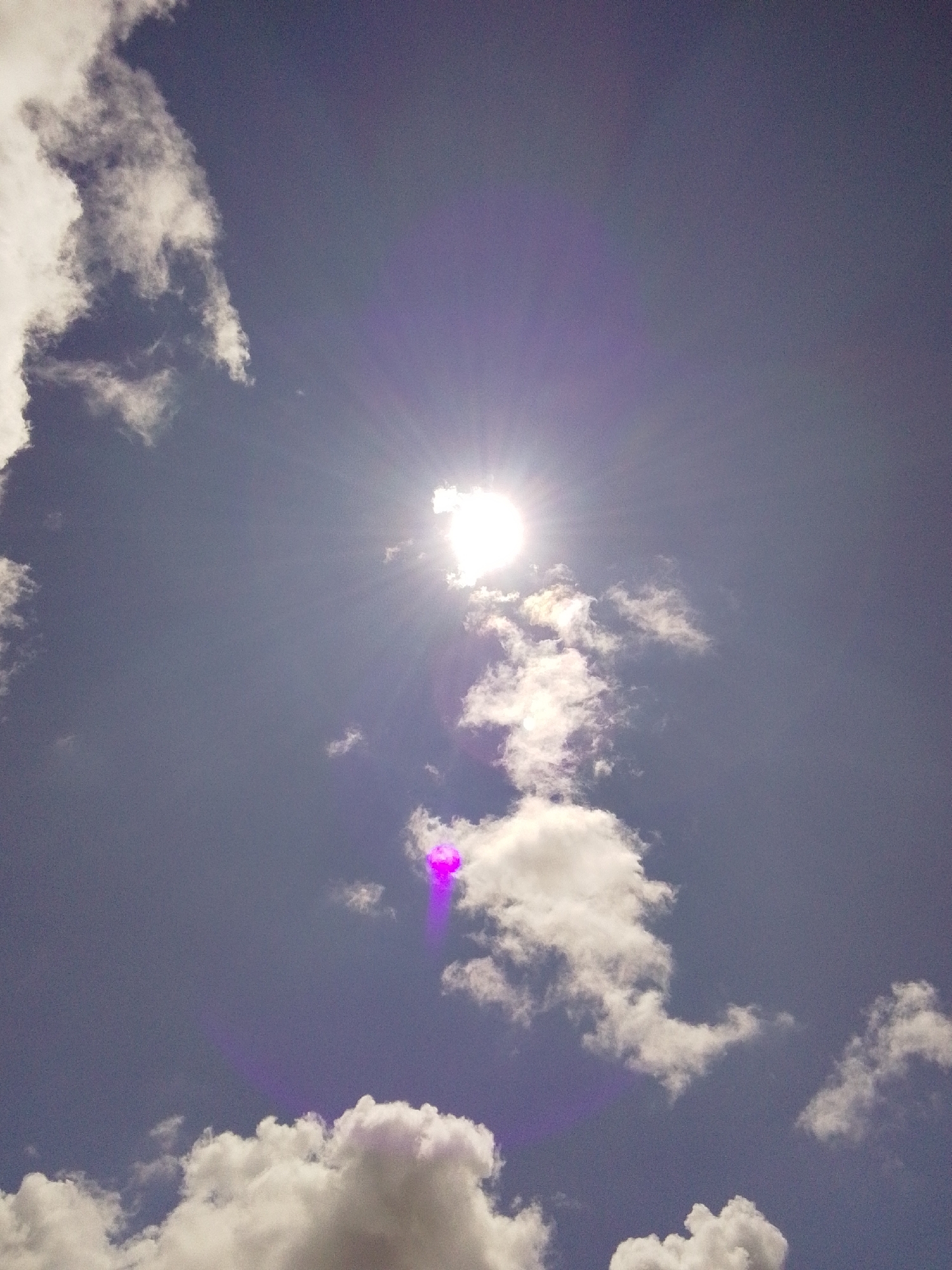 The image captures a daytime sky with scattered white clouds against a muted blue backdrop. The sun is partially visible in the upper left quadrant, emitting strong rays and creating a lens flare effect with purple and white hues. A distinct purple glow emanates from the clouds in the lower center. The scene appears to be an outdoor environment, characteristic of Serti, Nigeria, though no specific landmarks are identifiable. There are no people or man-made objects depicted in the image.