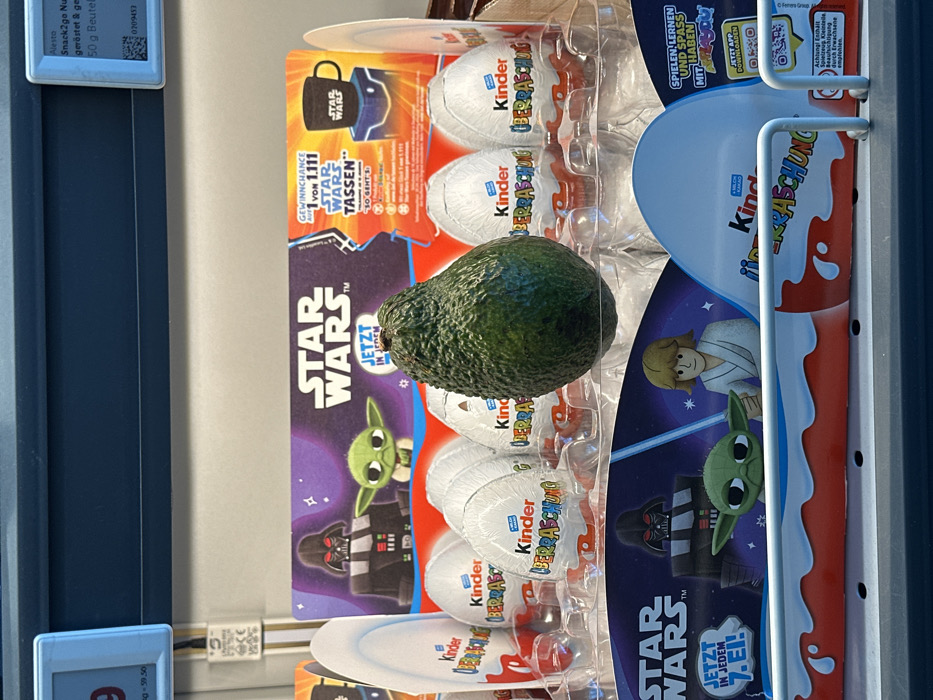 This image depicts a retail display in a store in Jestetten, Germany. The primary subject is a dark green, textured avocado centrally positioned within a clear plastic tray designed to hold confectionary eggs. Surrounding the avocado are multiple white, foil-wrapped Kinder Überraschung (Kinder Surprise) eggs, arranged upright in the tray's compartments.
Behind and beneath the eggs, Star Wars-themed promotional cardboard material is visible. The top banner features the "STAR WARS™" logo, text "JETZT IN JEDEM", and cartoon illustrations of Darth Vader and Grogu. It also advertises a "GEWINNCHANCE 1 von 1.111 STAR WARS TASSEN" (chance to win 1 of 1,111 Star Wars mugs), showing an image of a black Star Wars mug. The bottom banner similarly displays the "STAR WARS™" logo, text "JETZT IN JEDEM 7. EI!", and cartoon illustrations of Darth Vader, Grogu, and Luke Skywalker holding a blue lightsaber.
The environment includes a dark blue shelf or structure above the display. A white price tag on the left shows "9" and "q - 59.50". A smaller blue price tag on the right details "Alesto Snack2go Nu...", "geröstet & ge...", and "50 g Beutel", alongside a QR code and barcode. A white vertical element with "GS", "VDE", and "CE" markings is positioned between the price tags. The display rests on a lower shelf, partially obscured by a white metal guard rail in the foreground. No people are visible.