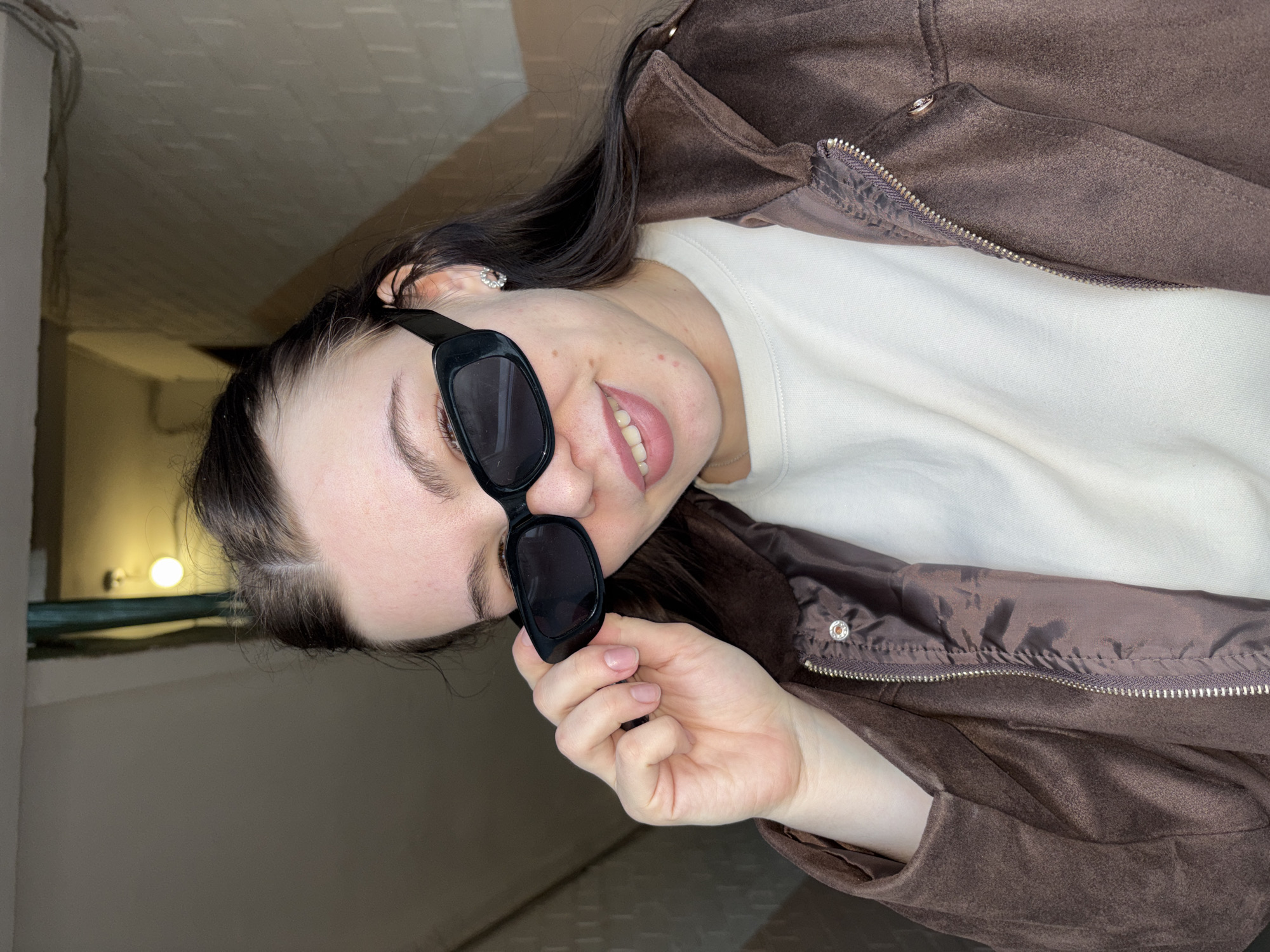 A young woman is captured in a close-up, chest-up shot, smiling broadly while wearing dark rectangular sunglasses. Her dark hair flows around her shoulders, and a small, sparkling earring is visible on her left ear. She is dressed in a dark brown, possibly suede or faux suede, zippered jacket, worn over a plain white long-sleeved top. Her right hand is positioned near her chin, gently holding the frame of her sunglasses. Her nails are natural and unpainted.
The background suggests an indoor, utilitarian setting, possibly a hallway or basement, with concrete-like walls and ceiling. The lighting is artificial and somewhat dim, with a warm-toned light fixture visible in the upper left of the frame. The overall atmosphere is casual. No text is visible in the image. The location context is Perm, Russia.