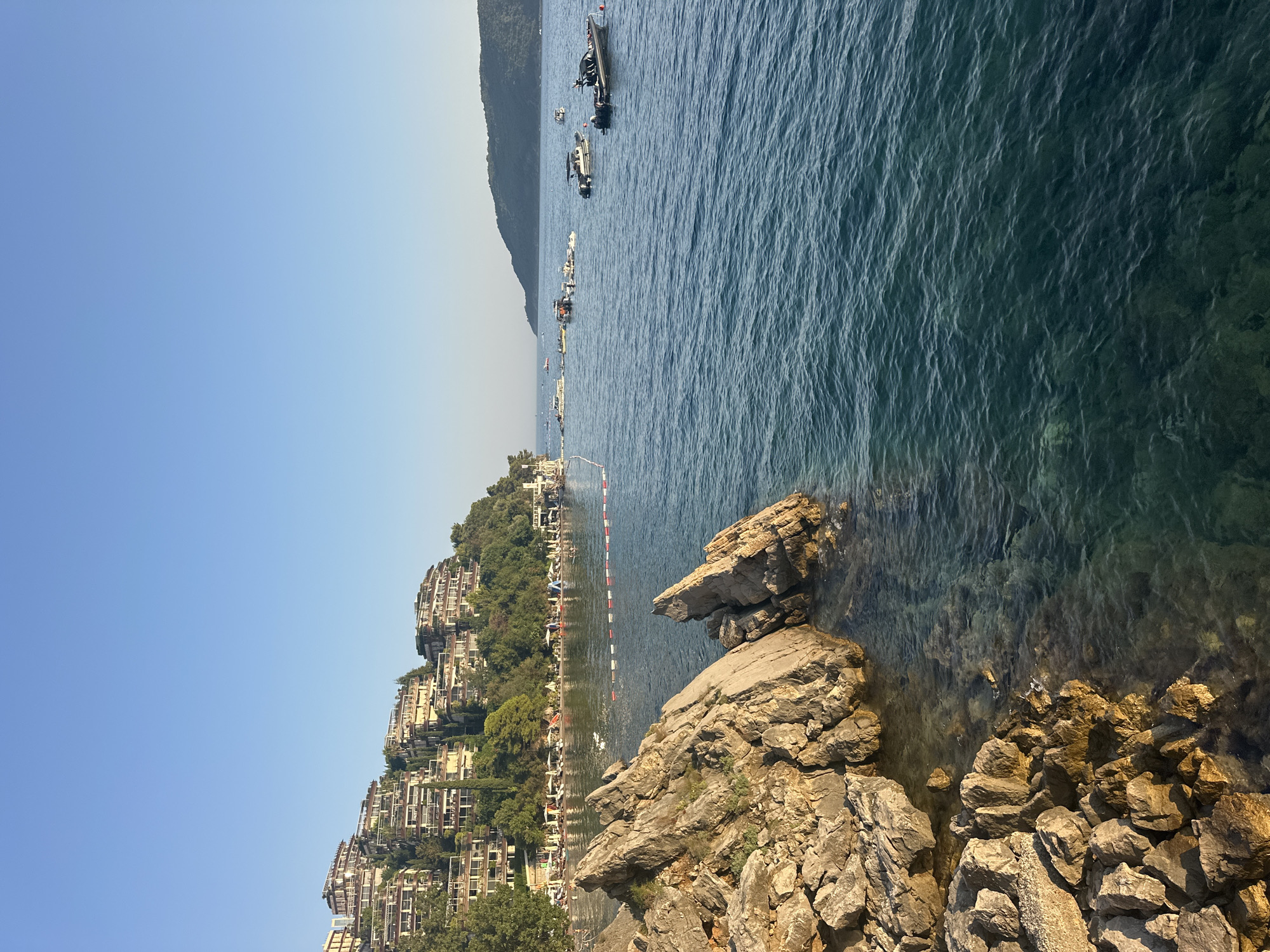 The image depicts a scenic view of the Adriatic Sea in Budva, Montenegro. In the foreground, rugged, sun-drenched rocks line the coastline, with clear turquoise water lapping at their base. Further out, the sea is a deeper blue, dotted with several boats and a floating barrier indicating a swimming area. Along the shore, a beach is visible with numerous umbrellas and lounge chairs, suggesting a resort or public recreational area. Dominating the hillside in the background are a series of terraced buildings with balconies, interspersed with lush green trees and vegetation. The sky above is a clear, vibrant blue, indicative of fair weather.