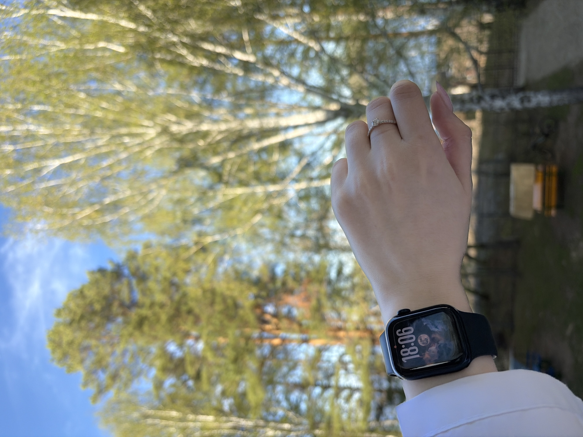 A person is wearing a wristwatch with a black band and a ring on their finger. The time displayed on the watch is 18:06. The background features trees and what appears to be a building. The image was captured in Chaikovsky, Russia.