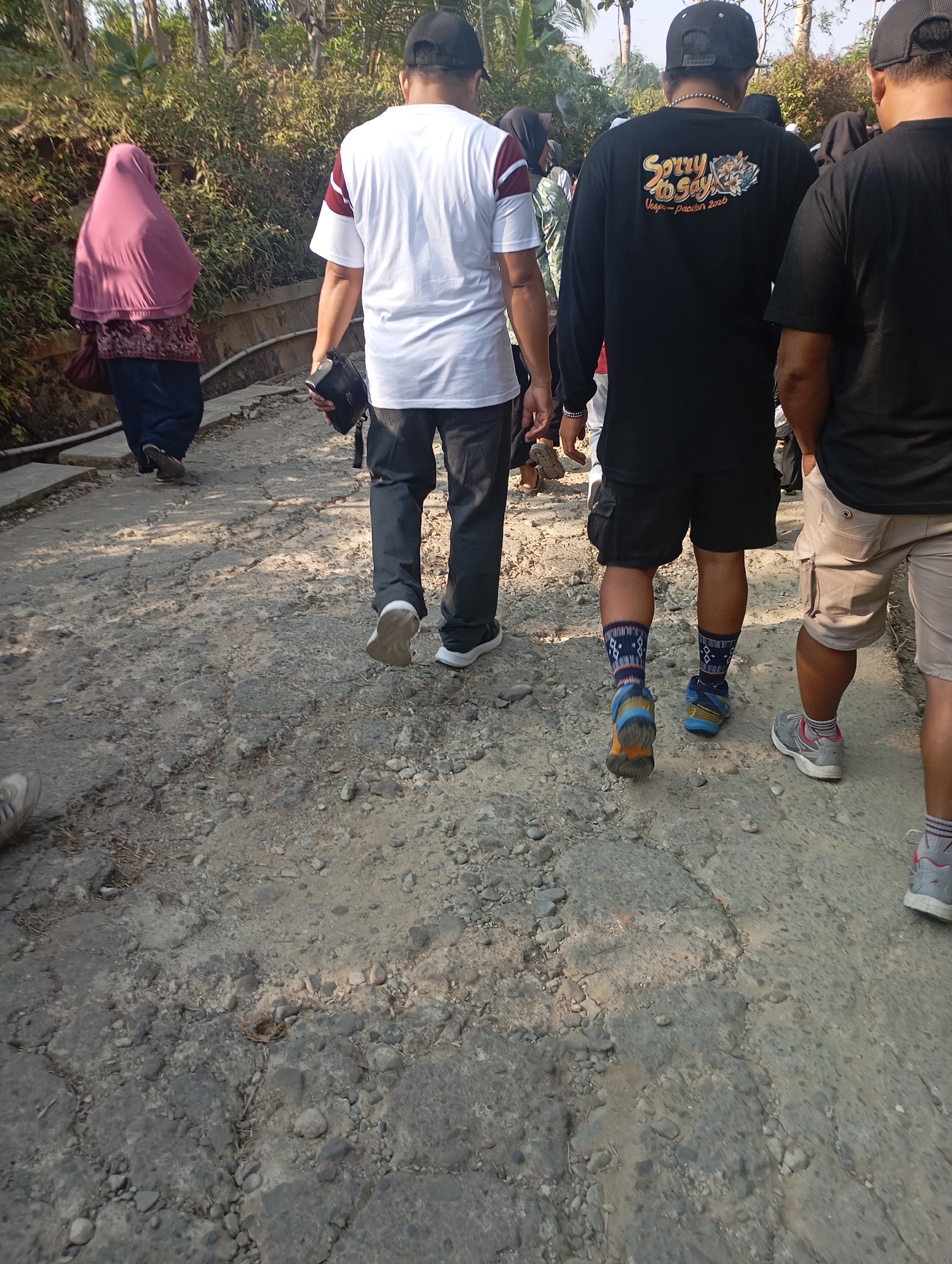In Ketepung, Indonesia, on a cracked, rocky path, a group of individuals walks away from the camera. The foreground is dominated by the rough texture of the path, with scattered stones and patches of dirt. Sunlight casts shadows and illuminates the scene.
To the left, a woman wearing a pink hijab and a patterned skirt walks ahead. She carries a dark bag in her left hand.
Following her, a man in a white t-shirt with burgundy stripes on the sleeves and dark pants walks, holding a black bag in his right hand.
Behind him, another man wears a black long-sleeved shirt with a graphic on the back that reads "SORRY TO SAY" and "Vespa-pacitan 2020". He is wearing black shorts and colorful socks with geometric patterns, and sneakers.
To the right, the legs of a person wearing light-colored cargo shorts and grey sneakers with pink accents are visible. Another person in a black t-shirt is partially visible further to the right. The background features lush green foliage and trees, suggesting an outdoor, possibly rural or park-like setting. The overall impression is of a casual outdoor activity or walk.