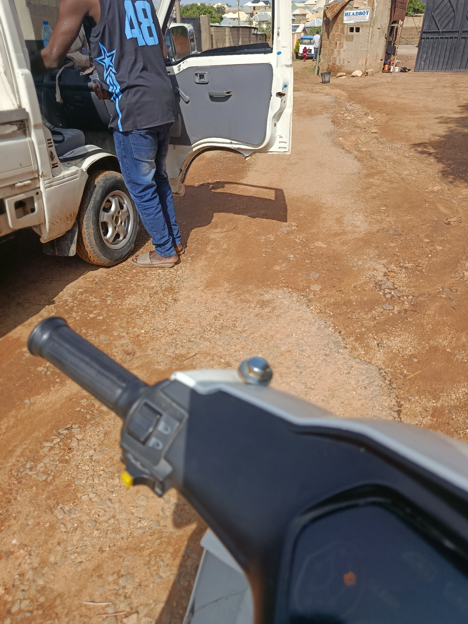 The image depicts a scene on a dirt road in Rafin Par, Nigeria, under bright daylight. In the foreground, the handlebars and part of the dashboard of a motorcycle are visible. The focus shifts to a white utility vehicle with its driver's side door open. A person, wearing a black tank top with blue "48" lettering and blue jeans, is partially entering or exiting the vehicle. The surrounding environment is characterized by dry, reddish-brown earth with scattered stones and gravel. In the background, a building with a sign that reads "HEADBOY" and more distant residential structures are visible. The overall impression is that of a functional, outdoor setting in a developing urban or semi-urban area.
