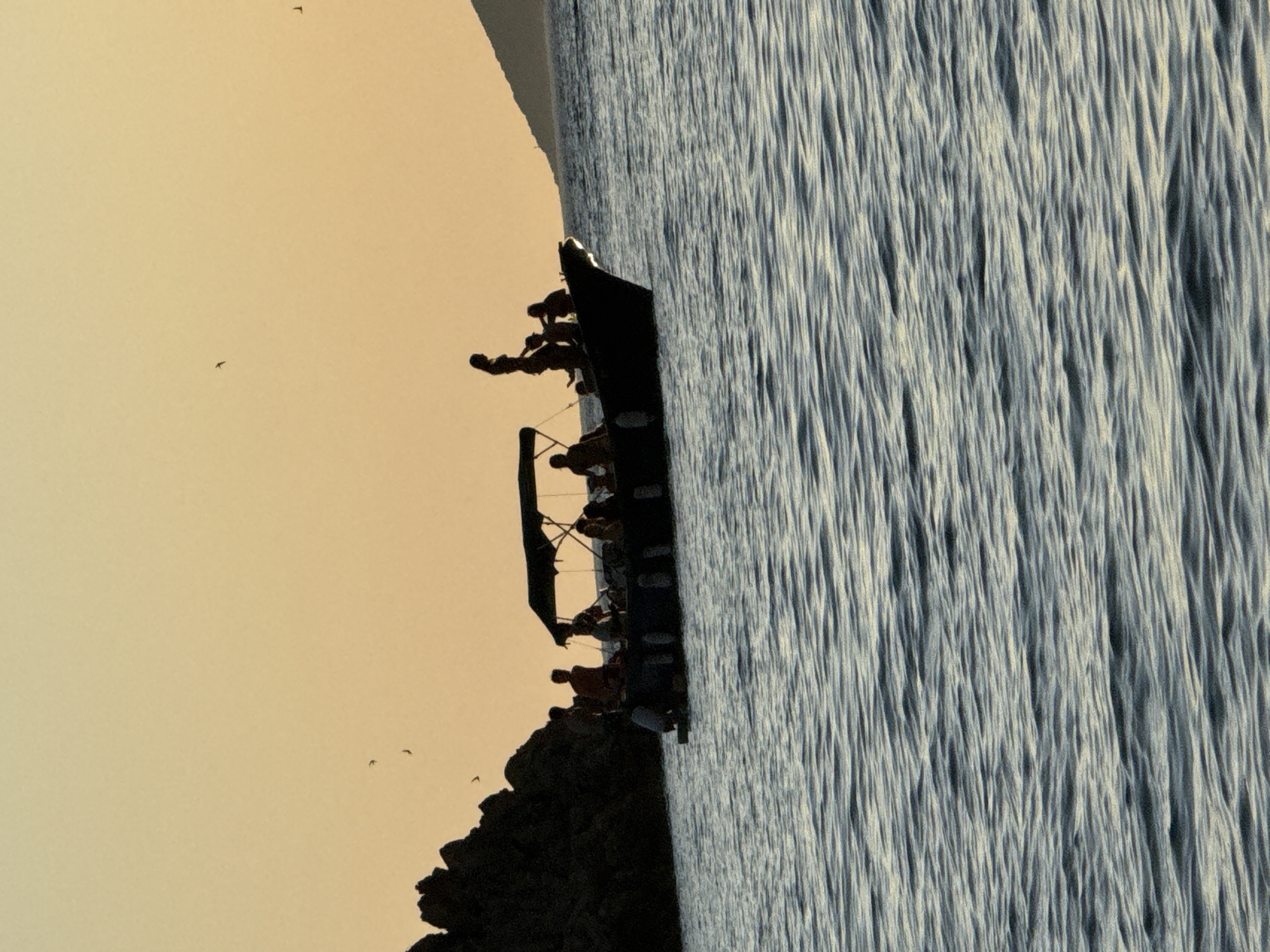 A silhouetted group of people are on a boat in the water near Sveti Stefan, Montenegro. The sky is a warm orange-yellow, indicating sunset or sunrise. The boat is dark and has a canopy. The people appear to be standing and sitting. To the left of the boat is a rocky, dark landmass. In the distance, to the right, is a faint outline of a hill with a structure on it. The water is choppy with visible waves.