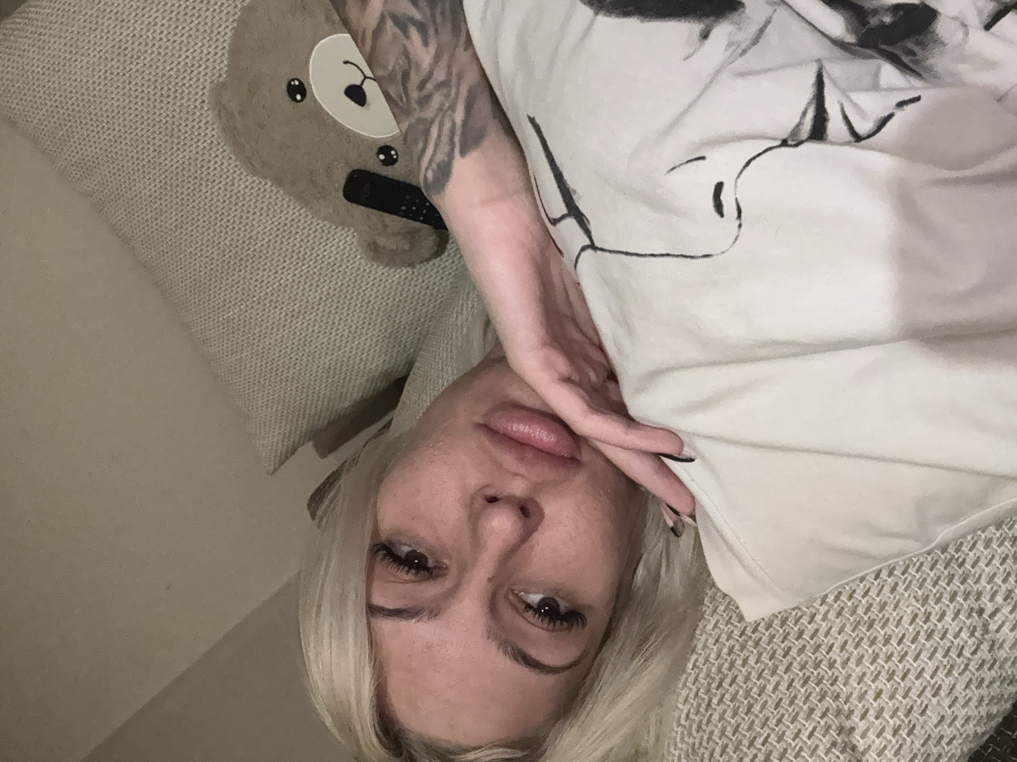 A person is lying down on a sofa or couch, with their head at the bottom of the frame. The person has light blonde hair and dark eyes. Their right hand is resting near their face, with dark nail polish visible. They are wearing a light-colored t-shirt with some sort of design on it. Beside them is a teddy bear-shaped pillow. The background consists of textured fabric of the sofa. The location is in an unknown city.