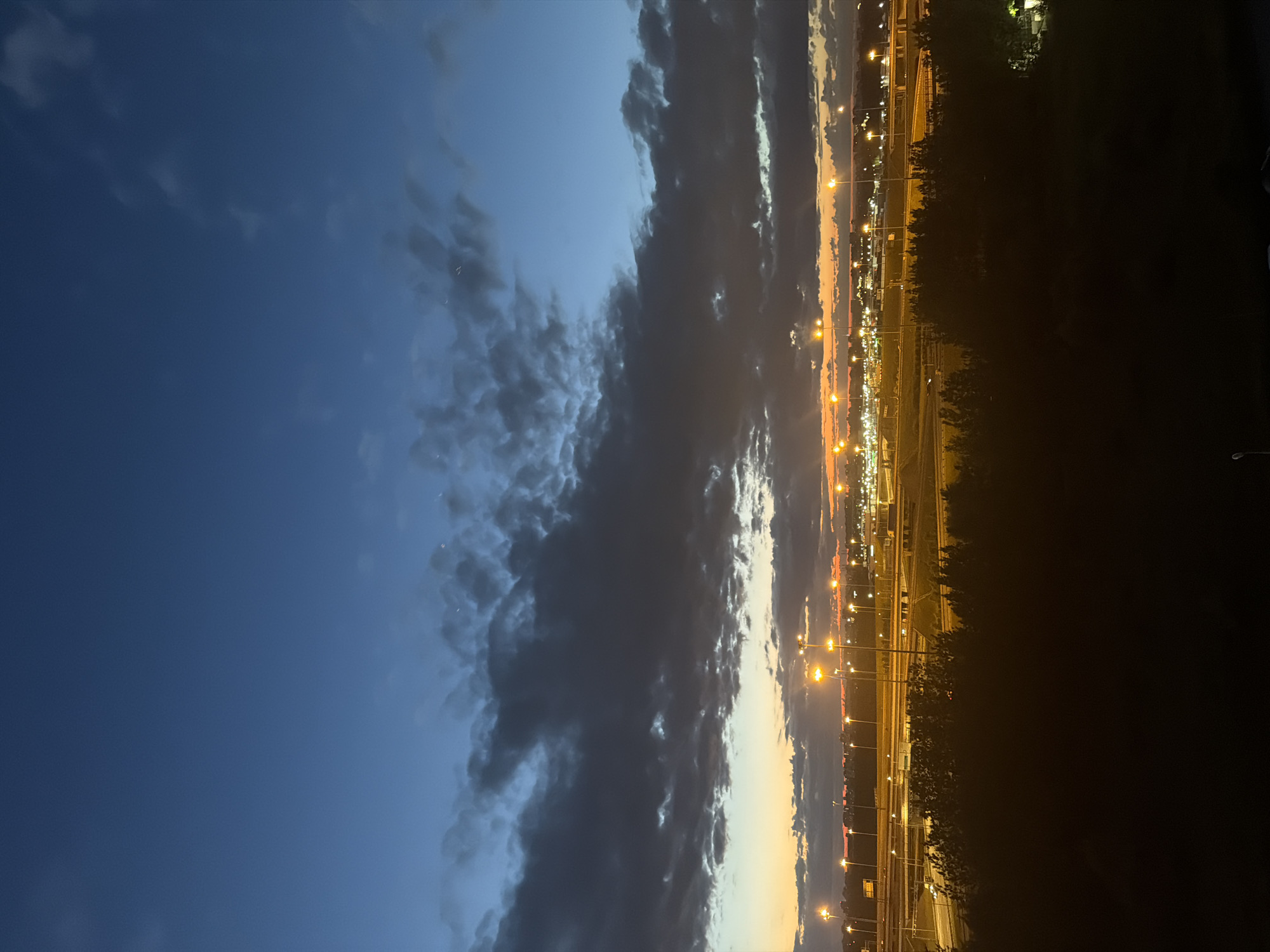 The image depicts a twilight scene over a city, with a dramatic sky transitioning from deep blue to orange hues near the horizon. Dark, textured clouds are scattered across the upper portion of the sky, interspersed with lighter patches. Below the clouds, a band of orange and yellow light illuminates the horizon, suggesting the last rays of sunset or the glow of city lights.
In the middle ground, a network of highways or roads is visible, marked by numerous streetlights casting a warm yellow glow. These lights are arranged in rows, illuminating the complex infrastructure of the roadways. In front of the highway, a line of dark, silhouetted trees creates a distinct foreground element, obscuring the immediate ground level. The overall impression is one of an urban landscape at the edge of day, with artificial light beginning to dominate as natural light fades.