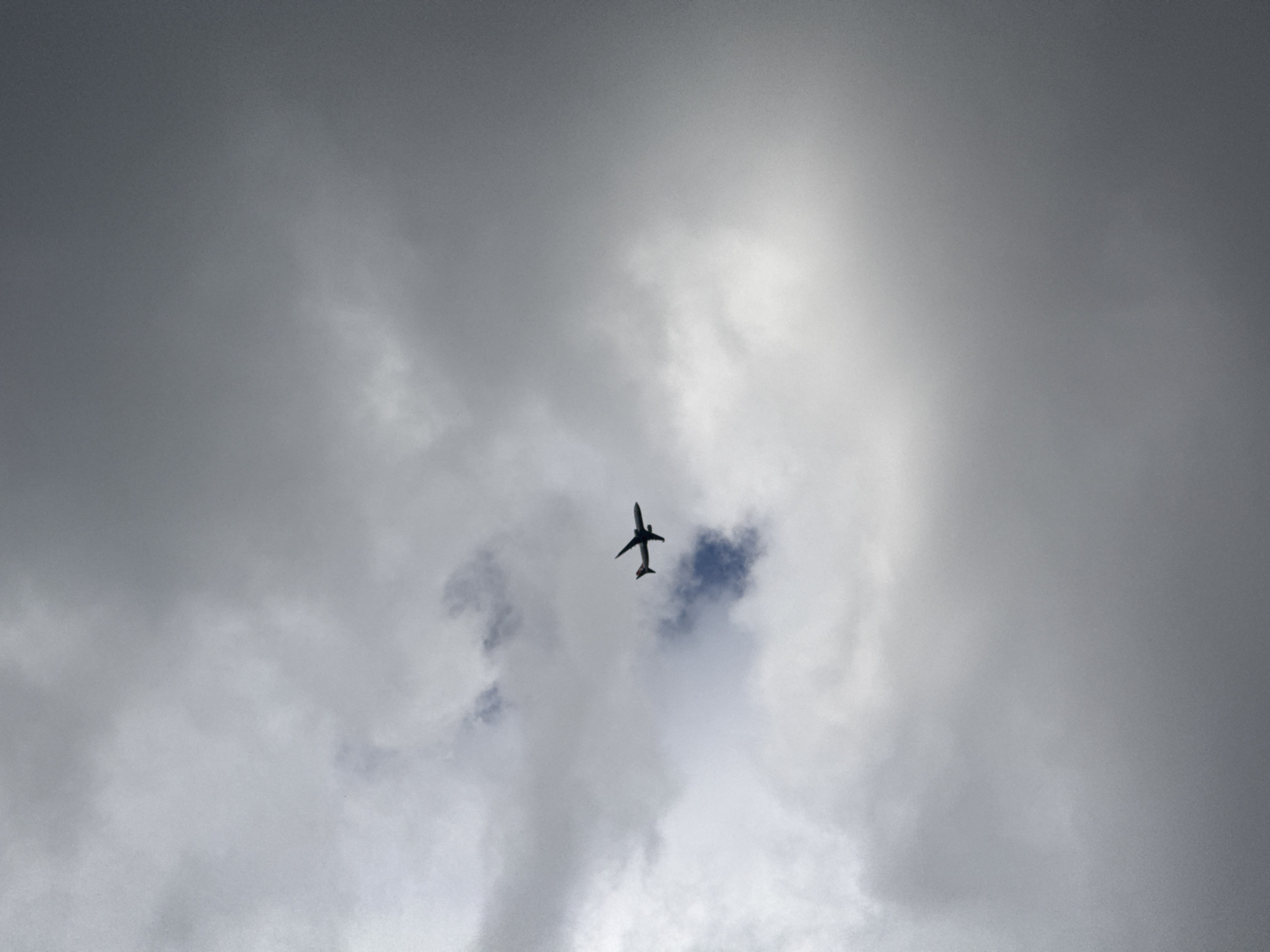A commercial airliner, appearing as a dark silhouette with a discernible red marking on its tailfin, is captured in flight against an overcast sky over Vila Nova de Gaia, Portugal. The aircraft is positioned in the upper mid-left portion of the frame, oriented with its nose directed towards the right. The sky consists of various formations of grey and white clouds, with a small, darker atmospheric patch visible below and slightly to the left of the aircraft. No other subjects or ground features are present, indicating a perspective directed upwards into the sky.