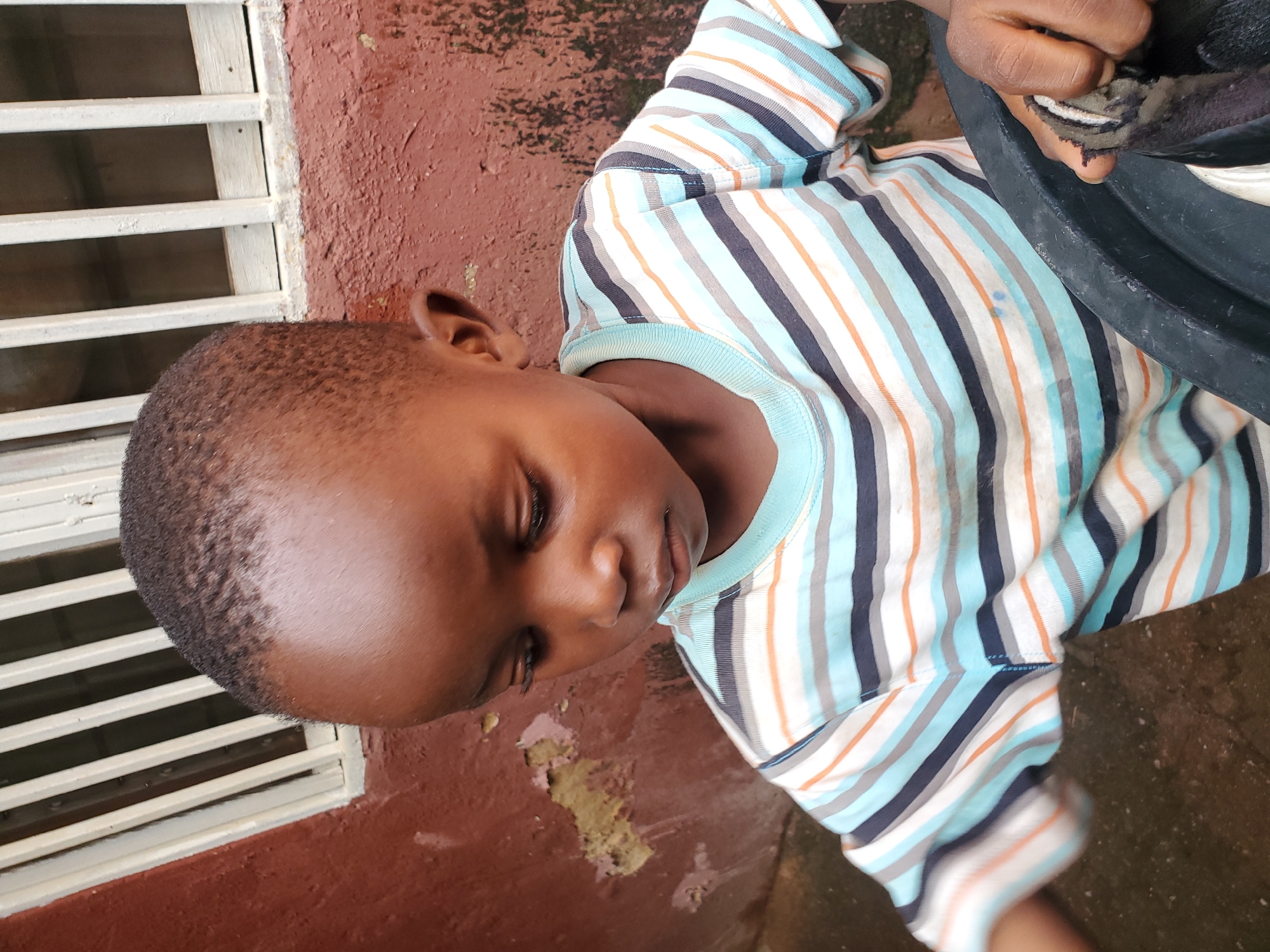 A close-up shot shows a young boy from the chest up, looking downwards and to his right. He has dark skin, short, dark hair, and is wearing a striped t-shirt with horizontal bands of light blue, white, gray, and dark blue, with thin orange stripes interspersed. The boy is positioned in front of a weathered, reddish-brown wall with patches of peeling paint and some green algae or moss. Behind him, a white barred window is visible. To the right of the boy, a portion of a black bucket or container is visible, and his right hand is reaching into it, holding a dark, patterned cloth. The ground appears to be damp and dirty concrete. This scene is set outdoors in a residential area of Jos, Nigeria.