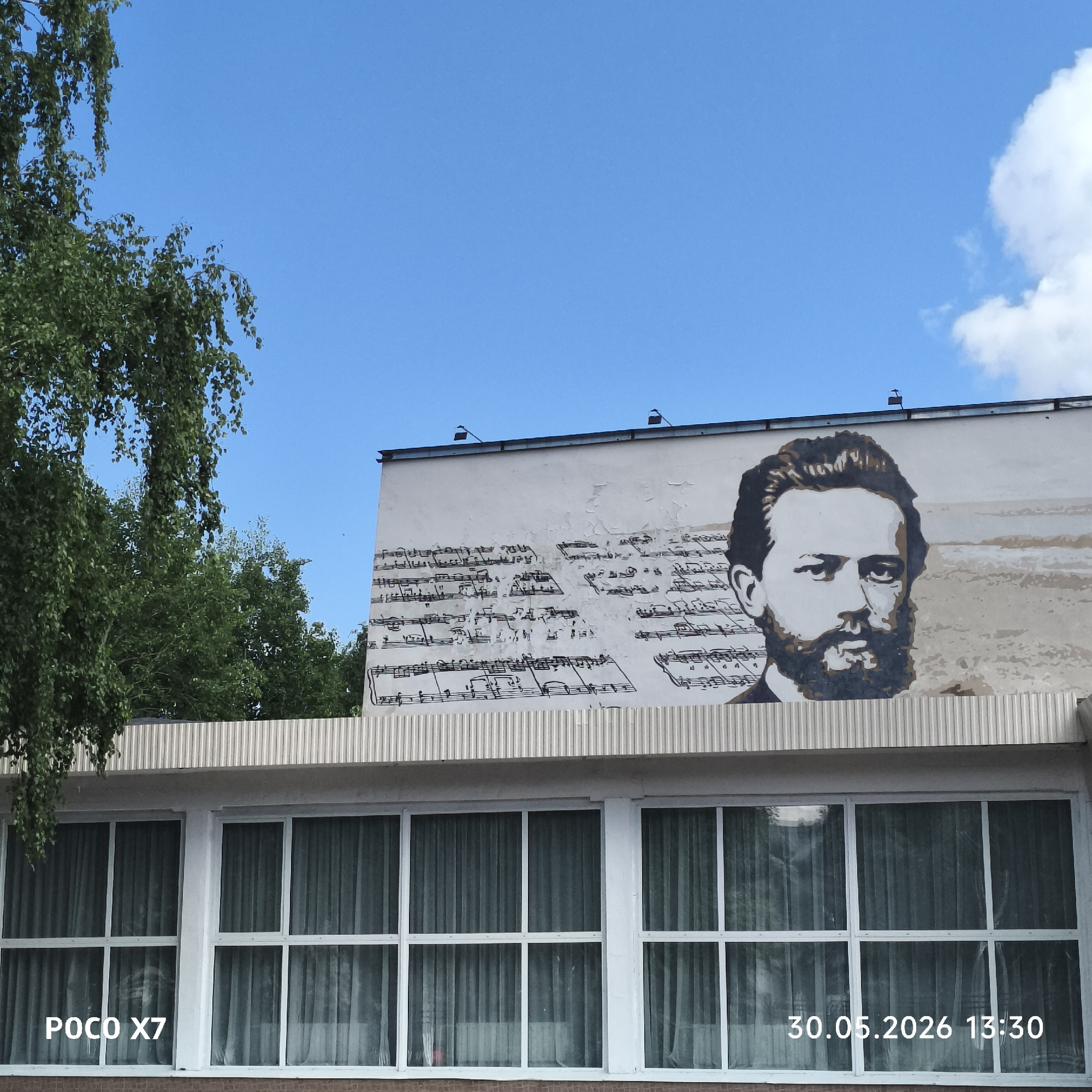 The image shows a portion of a building in the city of Chaikovsky, Russia. A mural on the upper part of the building depicts a portrait of a man and musical notation. The building has large windows with white frames. A tree with green foliage is visible to the left of the building. The sky is blue with some clouds. The date "30.05.2026 13:30" is printed in the lower right corner, and "POCO X7" is printed in the lower left corner.