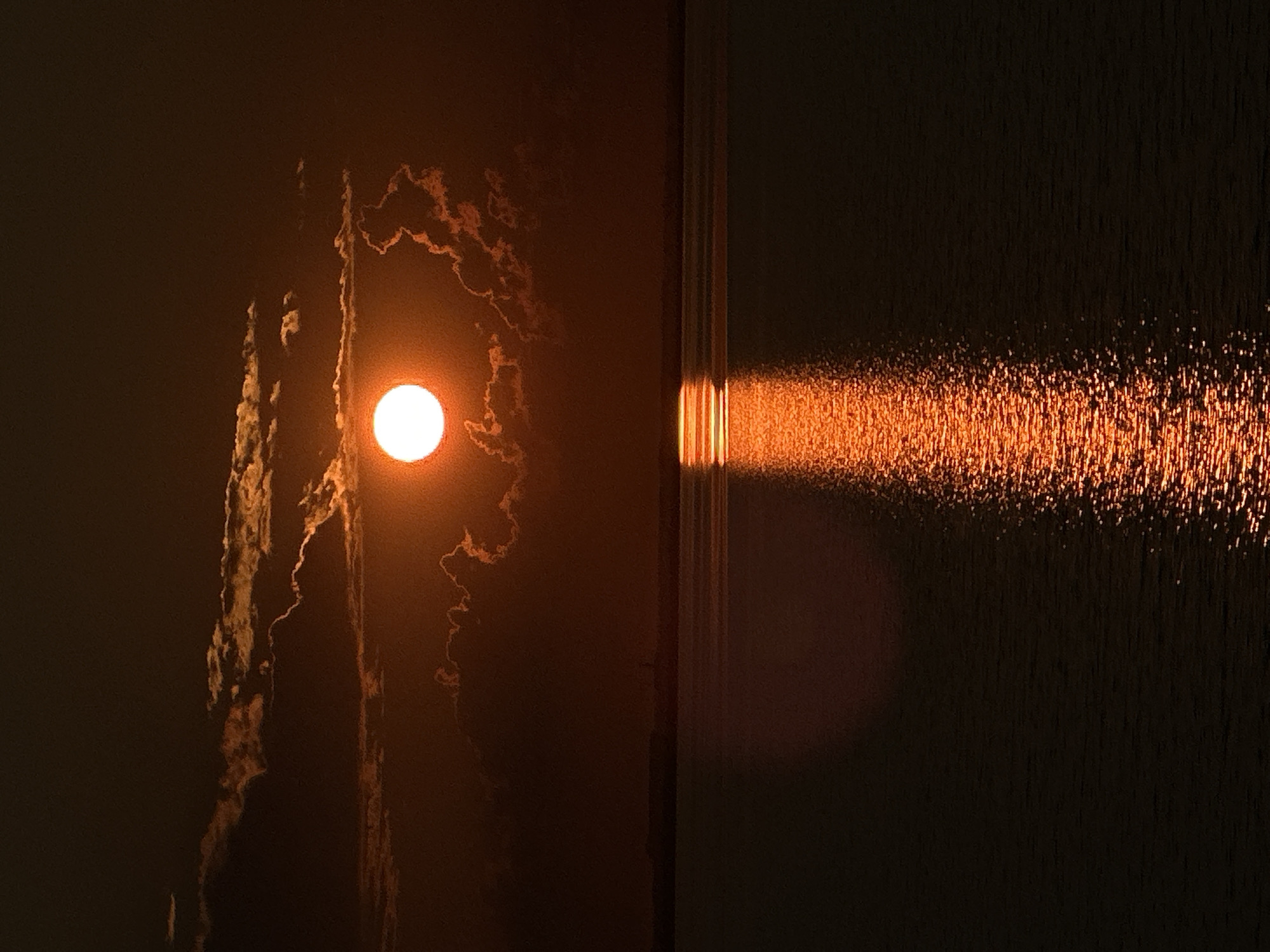 The image captures a sunset viewed through a window, with the sun appearing as a bright, glowing orb partially obscured by wispy, illuminated clouds. The light from the setting sun casts a warm, orange hue across the scene. On the left side of the image, the sun is centered, with the clouds creating abstract, textured patterns around it. On the right, the sunlight streams through a vertical window frame, creating a bright, shimmering reflection on a textured surface, likely water or a similar reflective material. This reflection is elongated and appears to be moving or rippling, suggesting a sense of dynamism. The overall mood is serene and contemplative, evoking the beauty of a natural phenomenon observed from an indoor setting. The lighting suggests either late afternoon or early evening, as the sun is low on the horizon. There is no visible text in the image. The location context is Kokshetau, Kazakhstan.