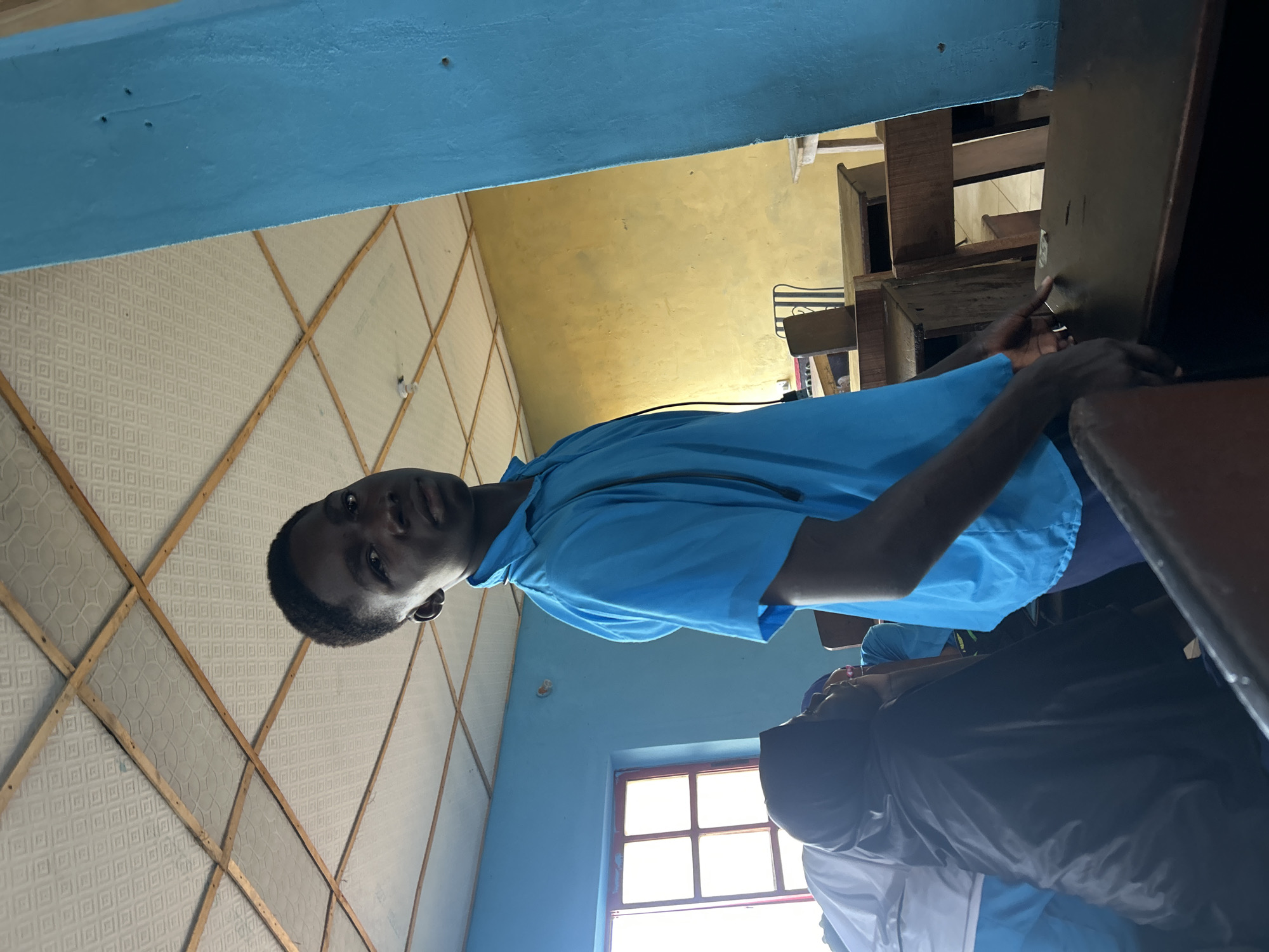 The image depicts an indoor scene, likely a classroom in Ikorodu, Nigeria, with several individuals present. The primary subject is a young person with dark skin and short dark hair, positioned centrally and looking directly at the camera. They are wearing a light blue, short-sleeved collared shirt, suggesting a school uniform.
Behind and to the right of the main subject, another individual in a similar light blue shirt is partially visible. In the lower right corner, a person is seen from behind, wearing a black head covering and a black long-sleeved top over a white garment.
The room features light blue walls in the foreground, transitioning to a yellowish-cream color further back. The ceiling is constructed with textured, square panels supported by a wooden grid framework. Wooden classroom desks or benches are visible throughout the space, reinforcing the educational setting. A multi-paned window on the right side allows bright daylight to enter, indicating it is daytime. A bare light bulb hangs from the ceiling in one section. No specific activity beyond being present in the classroom is clearly depicted, and there is no visible text.