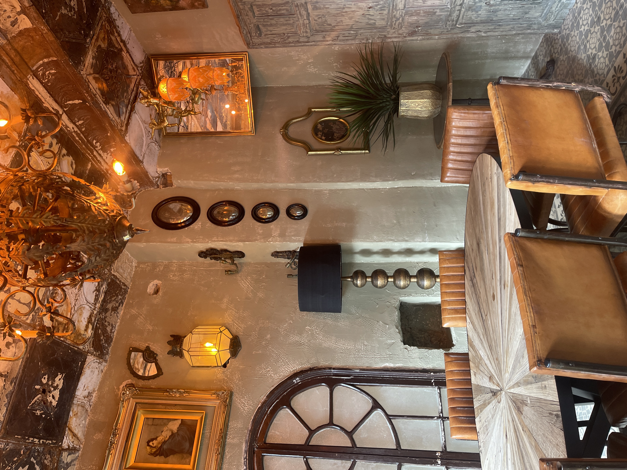The image depicts an interior view in Jerez De La Frontera, Spain. The setting appears to be within a restaurant or bar, indicated by the presence of tables and leather-upholstered chairs. The decor is eclectic, including an antique chandelier, framed paintings, and ornamental wall decorations. Architectural elements include a segmented arched window and a textured ceiling. The lighting is a mix of natural and artificial, with lamps and a window providing illumination.