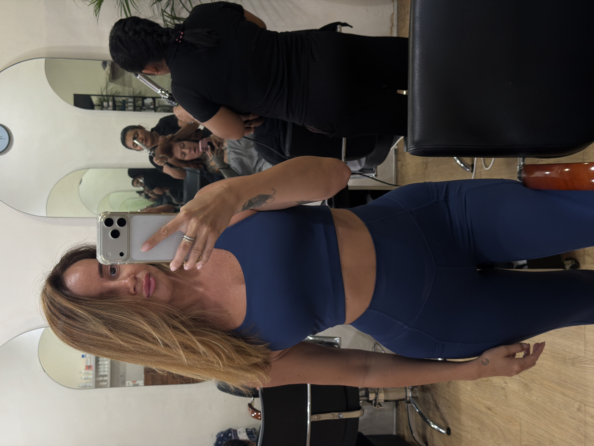 In a beauty salon in North Kuta, Indonesia, a woman takes a selfie in a mirror. She is wearing a dark blue sports bra and leggings. Her light brown hair is styled loosely. In the mirror's reflection, two other people are visible. One is a woman with dark hair in a braid, wearing a black shirt, holding a curling iron, and working on another person's hair. The other is a woman with red hair who is looking at her phone. The salon has light-colored walls, arched mirrors, and wooden floors. There are salon chairs and shelves with products.