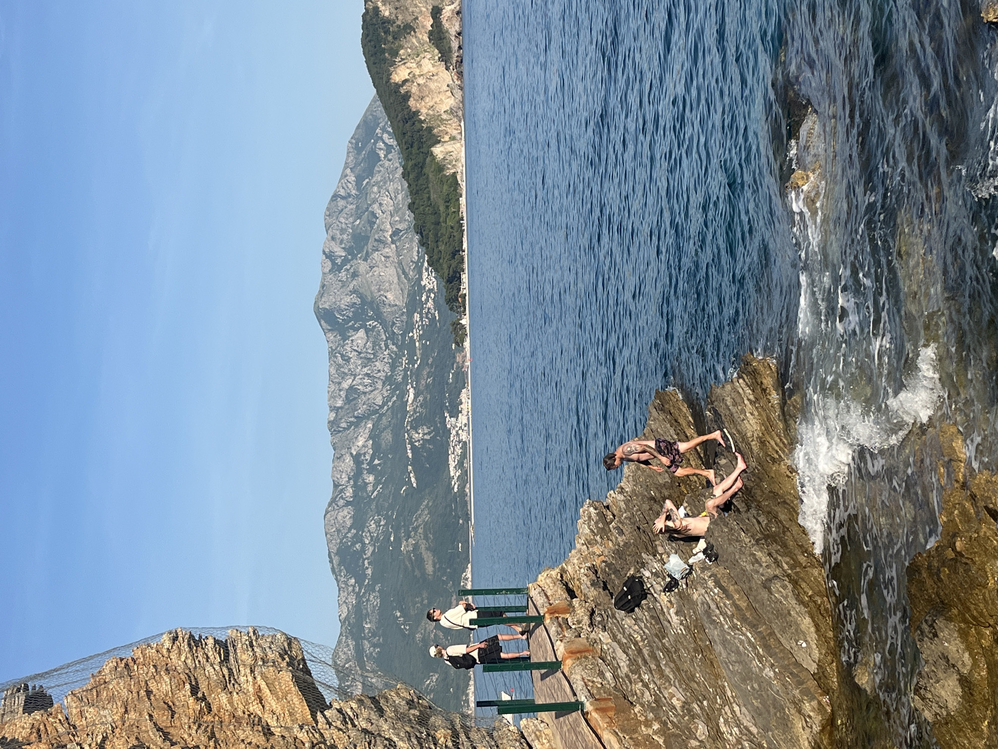 This panoramic photograph captures a vibrant coastal scene in Budva, Montenegro, on a clear, sunny day. The deep blue sea, with gentle ripples and white foamy waves crashing against the rocks in the foreground, dominates the right side of the frame. On the left, majestic, rugged mountains, partially covered in greenery, rise dramatically, with a coastal town featuring white and light-colored buildings nestled at their base along the distant shoreline.
In the mid-ground and foreground, a group of people is seen enjoying the rocky coastline. Three individuals stand on a wooden walkway with green railings built into the cliff face, observing the expansive view. One person wears a light shirt and sunglasses, another a dark shirt and backpack, and a third a white cap. Closer to the water's edge, on a natural rocky outcrop, three more people are present. One man with tattoos, wearing dark patterned shorts, stands poised on the very edge of the rocks, seemingly preparing to enter the water or enjoying the view from a prime spot. Nearby, another man, also with visible tattoos and wearing light shorts, is lying relaxed on the sun-warmed rocks with his arms bent over his head. A third person is partially visible lying or sitting near him. A black backpack and some light clothing are placed on the rocks beside them. The rocks themselves are a mix of dark brown and grey, exhibiting a layered, jagged texture. A protective wire mesh is visible along the top of the cliff on the far left, suggesting rockfall mitigation. The overall atmosphere is one of leisure and appreciation for the beautiful natural surroundings. There is no visible text in the image.