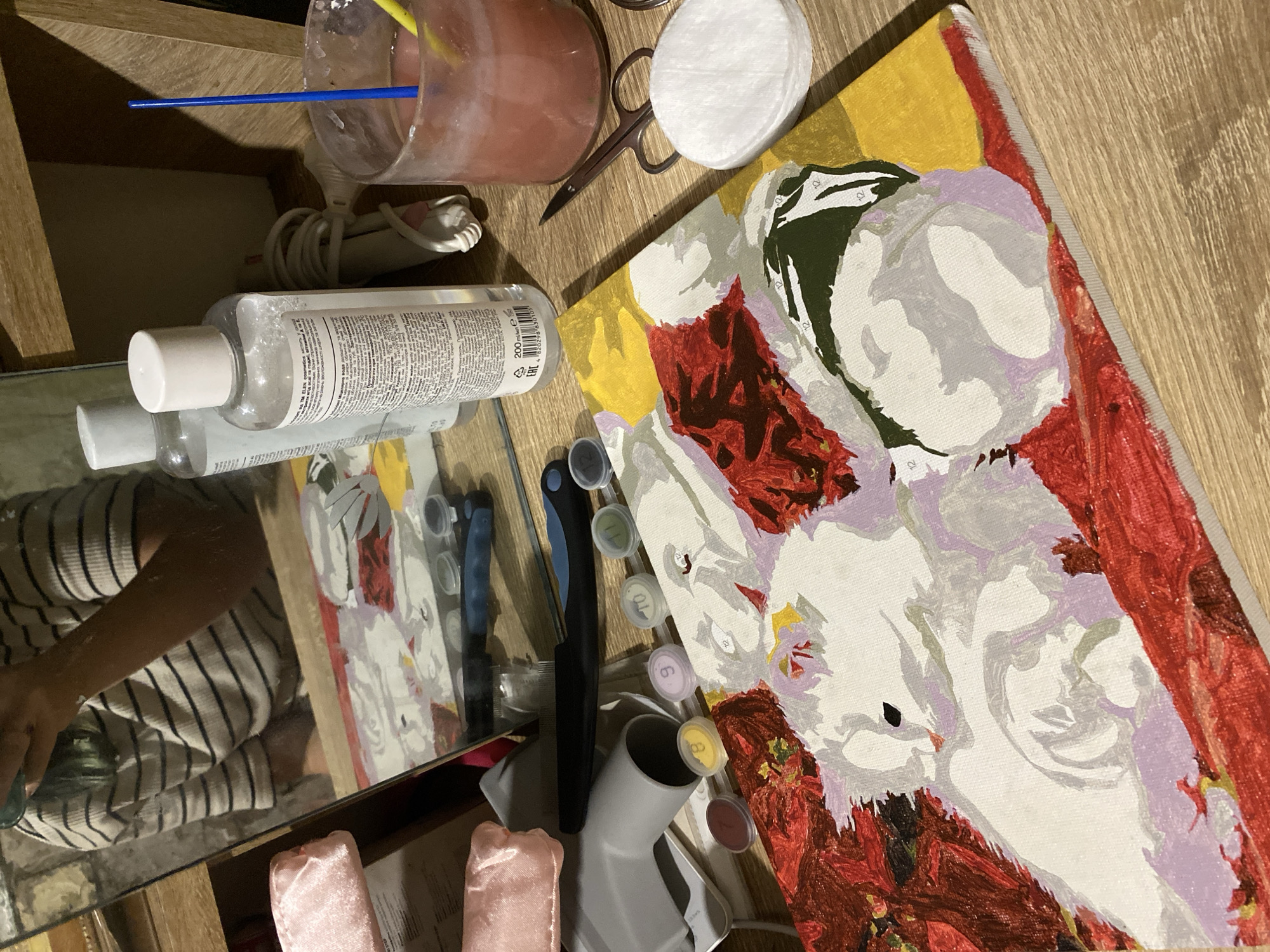 The image shows a partially completed paint-by-numbers artwork depicting a white cat nestled amongst red and yellow foliage. The canvas rests on a wooden surface, likely a desk or table. Scattered around the canvas are various art supplies, including small pots of paint with numbers, a paintbrush, a container of pink liquid, scissors, cotton pads, and a bottle of micellar water. In the background, a mirror reflects a person wearing a striped shirt, suggesting the artwork is being created by someone. The scene appears to be an indoor setting, possibly a home environment. There is no visual information in the image to indicate a location in Nova Praha, Ukraine.