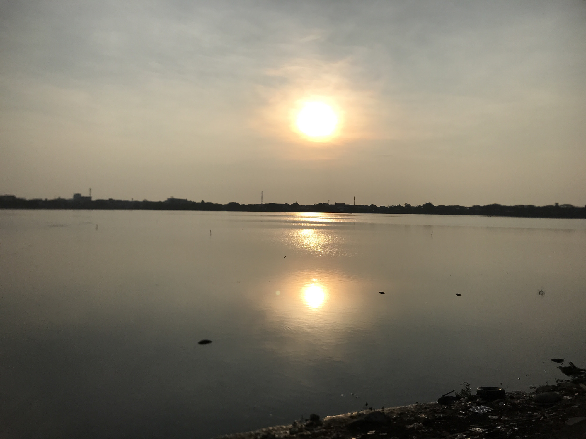 The image depicts a body of water with the sun setting or rising in the background. The sun's reflection is visible on the water's surface. In the distance, a cityscape or line of trees is silhouetted against the horizon. The foreground shows the shoreline or edge of the water, with some debris or rocks visible.
Specific details consistent with Lhokseumawe, Indonesia, are not definitively discernible from the image alone. However, the presence of a large body of water, a coastal city skyline, and the tropical lighting could align with the general characteristics of Lhokseumawe. Without further information or specific landmarks, pinpointing the location within the city is not possible.