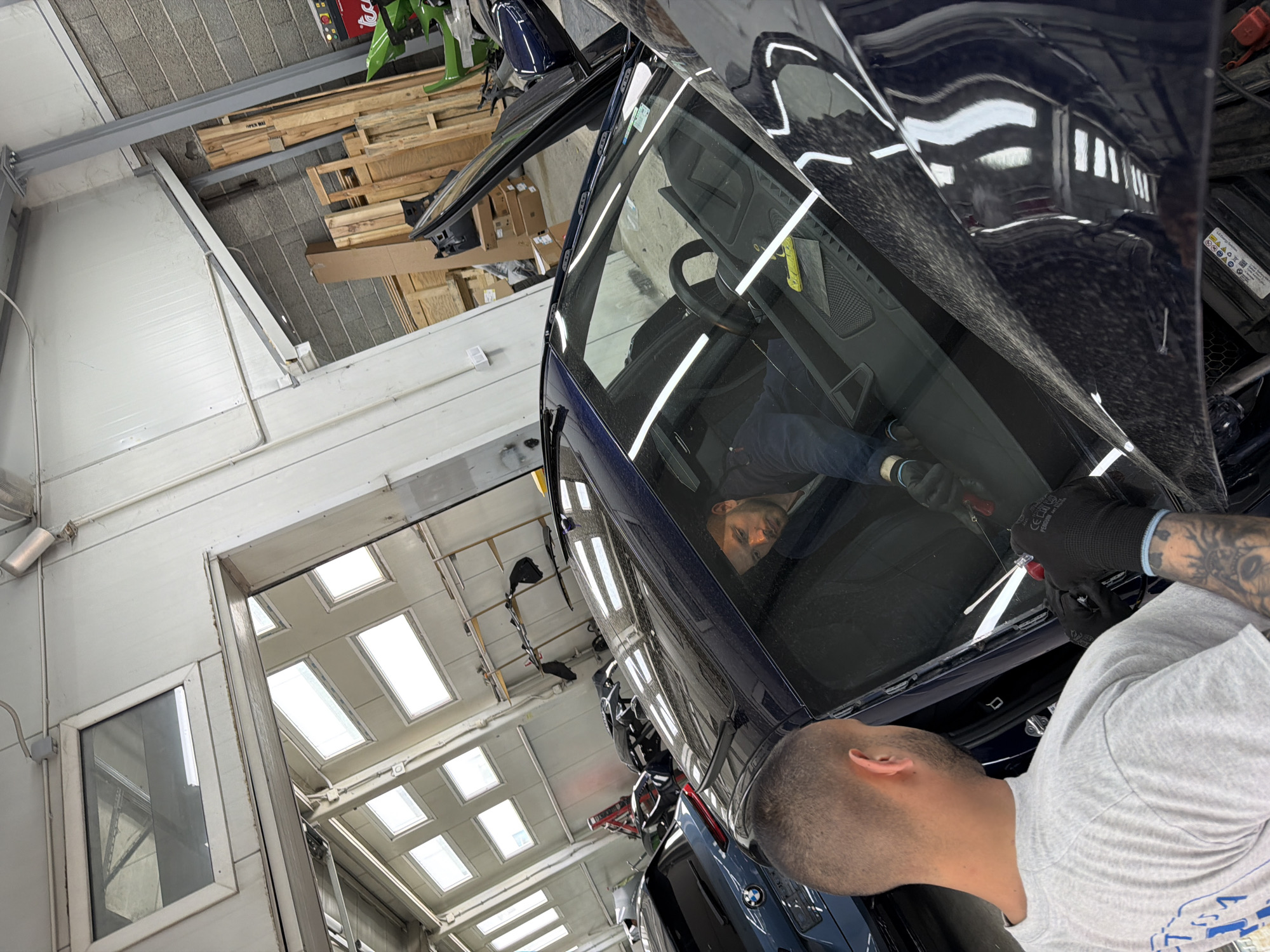 The image shows two men working on a blue BMW in what appears to be an auto repair shop in Burgas, Bulgaria. The angle of the photo makes it hard to see details, but it's clear that the men are working on the car's windshield. One man is reflected in the windshield, holding a tool. The other man, with short hair and a gray shirt, is seen from the neck up, looking at the car. He has tattoos on his arm. The background shows a typical industrial space with bright overhead lighting, a white wall with windows, and wooden pallets stacked up near the ceiling. The overall impression is that the image was taken inside a car repair or body shop.