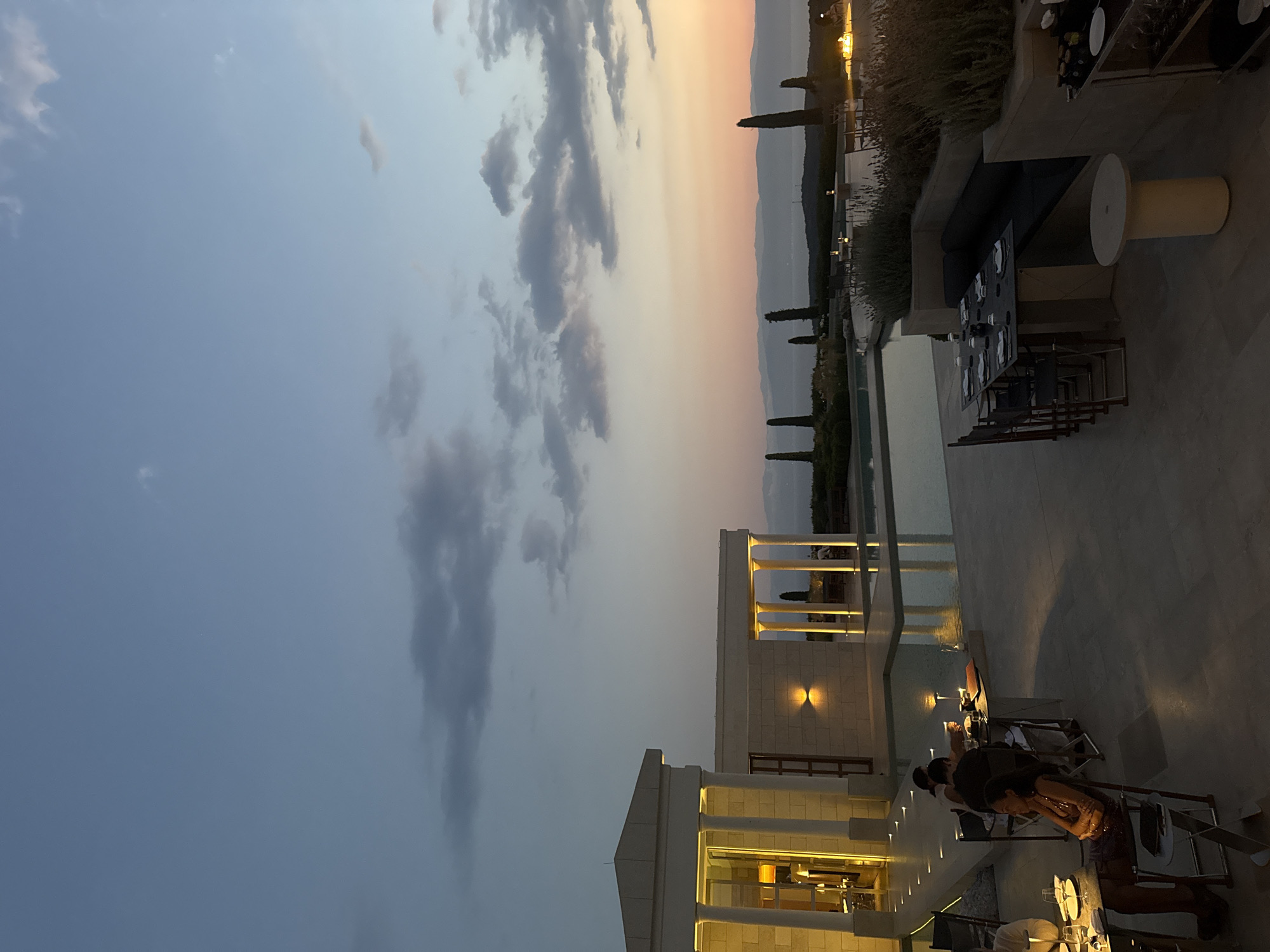 The image shows an outdoor dining area at dusk in the Municipal Unit of Kranidi, Greece. A sleek infinity pool with a modern architectural structure featuring classical columns is visible to the left, illuminated by warm interior lighting. Several tables are set for dining, with plates, glasses, and cutlery arranged on them. Diners are seated at some of the tables, with one woman in a sparkly dress in the foreground appearing to be dining alone. In the background, a serene landscape with distant mountains and a body of water can be seen under a twilight sky with scattered clouds. Tall, slender trees punctuate the horizon. The overall atmosphere suggests a luxurious and tranquil dining experience.