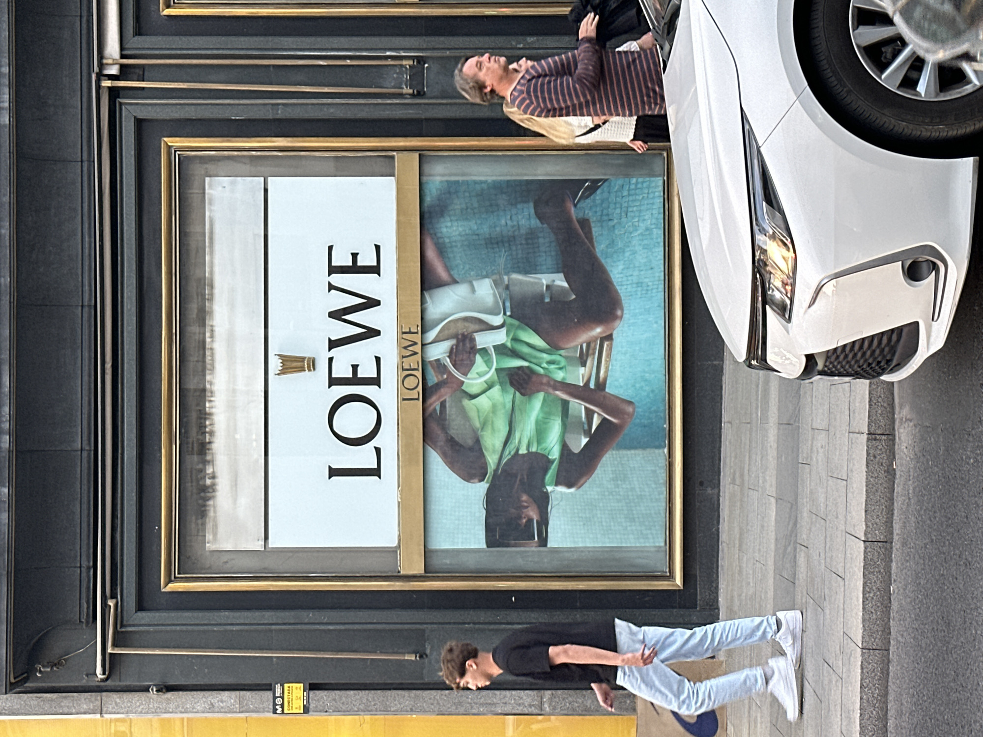 The image captures an exterior scene in Madrid, Spain, featuring a Loewe store window. The window displays a Loewe advertisement with the brand's name prominently featured alongside a model in a green top and holding a white bag, sitting in front of what appears to be a pool. A middle-aged person wearing a striped shirt is visible in the upper portion of the reflection on the glass of the window. A white car is visible on the right. Below the window, a young man dressed in a black T-shirt and light blue jeans walks on the sidewalk. The environment includes the store façade, sidewalk, and a portion of the street.