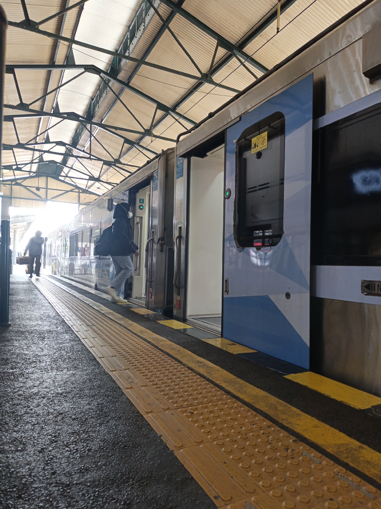 The image depicts a train station platform in Klaten, Indonesia, with a train stopped at the platform. The platform has a textured yellow tactile paving strip running along its edge, separating it from the dark asphalt. The train is modern, with a light blue and white geometric design on its side. One of the doors is open, and a person wearing a dark hooded jacket, light-colored pants, and a white face mask is stepping onto the train. This individual is carrying a backpack. In the background, another person is walking away from the train, carrying a rectangular bag. The station has a covered roof supported by metal beams, allowing natural light to filter through. The overall atmosphere suggests a typical scene at a public transportation hub.